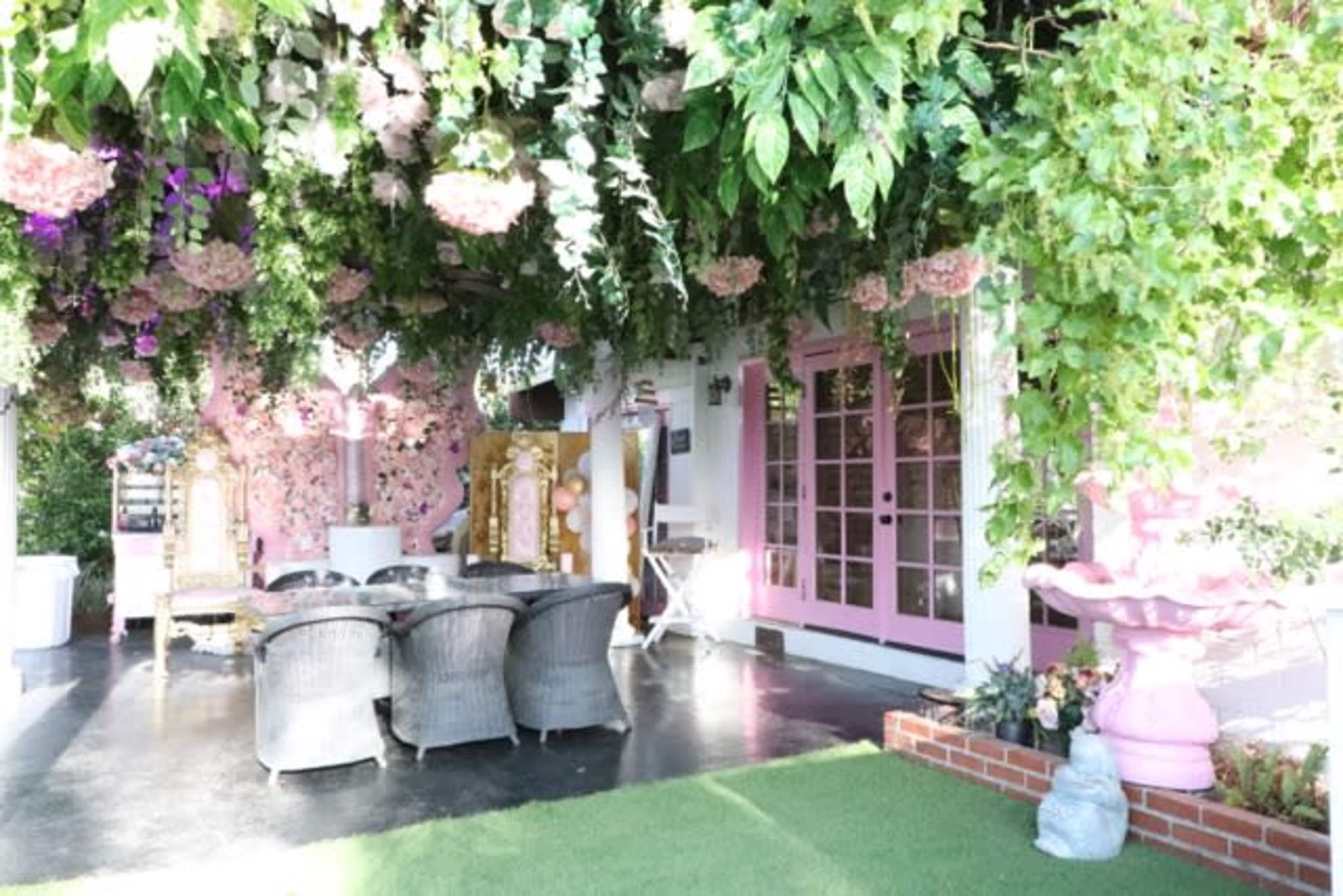 A decorated outdoor space features a canopy of greenery and flowers above a circular dining table surrounded by wicker chairs.