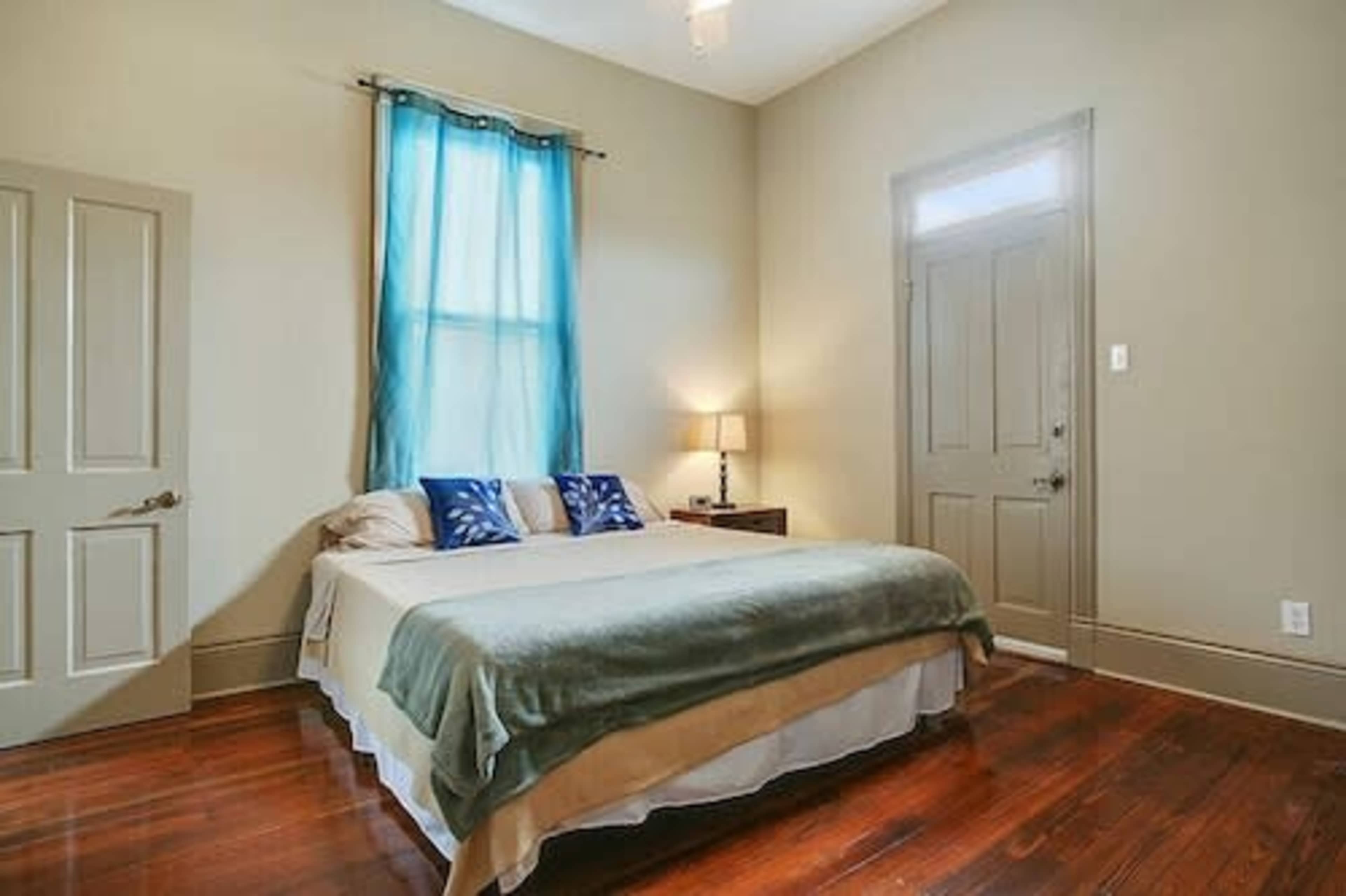 A well-lit bedroom with a bed covered in a light blanket, a bedside table with a lamp, and a window dressed with blue curtains.
