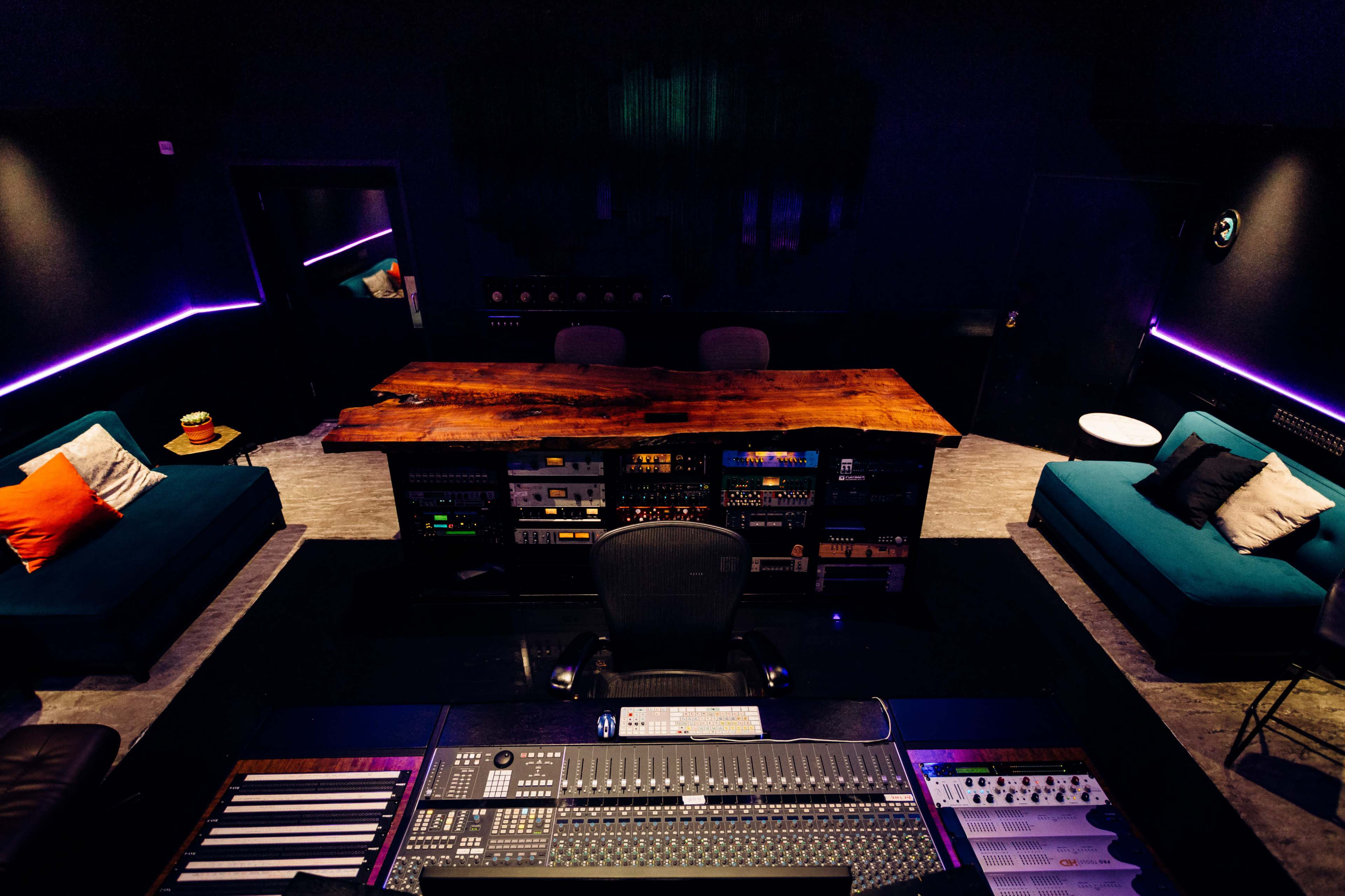 A modern recording studio featuring a large wooden table, a central mixing console, and teal couches arranged around the space.