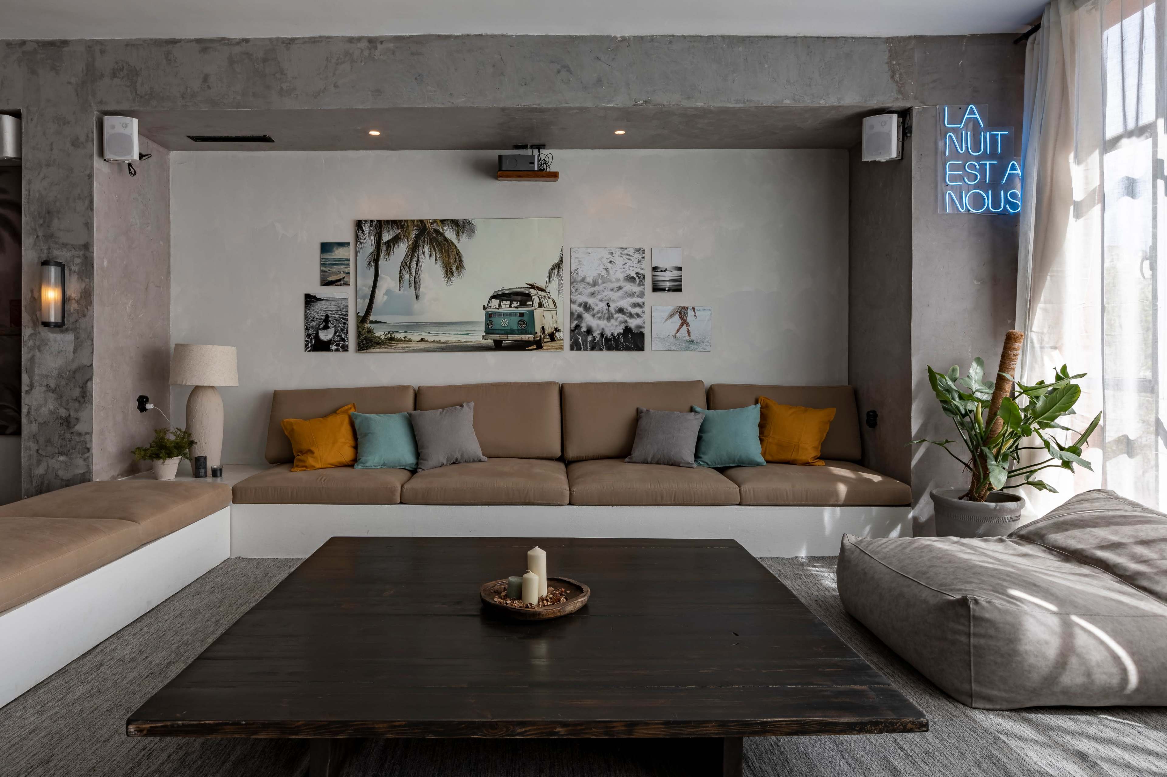 A modern living room with a large beige sectional couch, a dark wooden coffee table, and tropical-themed wall art, featuring a plant and a lamp in the background.