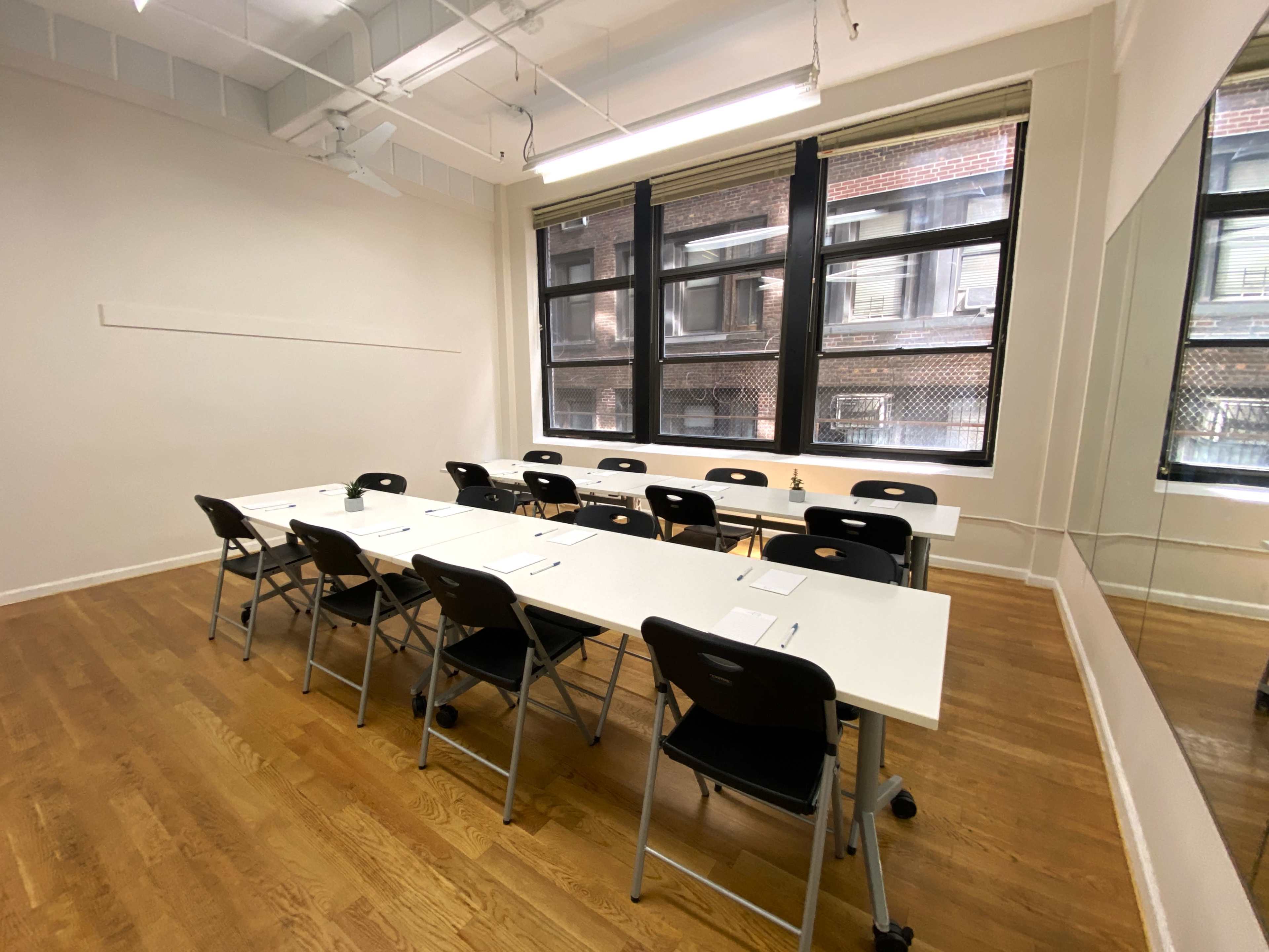 A bright, minimalist classroom features several foldable tables arranged in a U-shape with black chairs, alongside large windows and a mirror.
