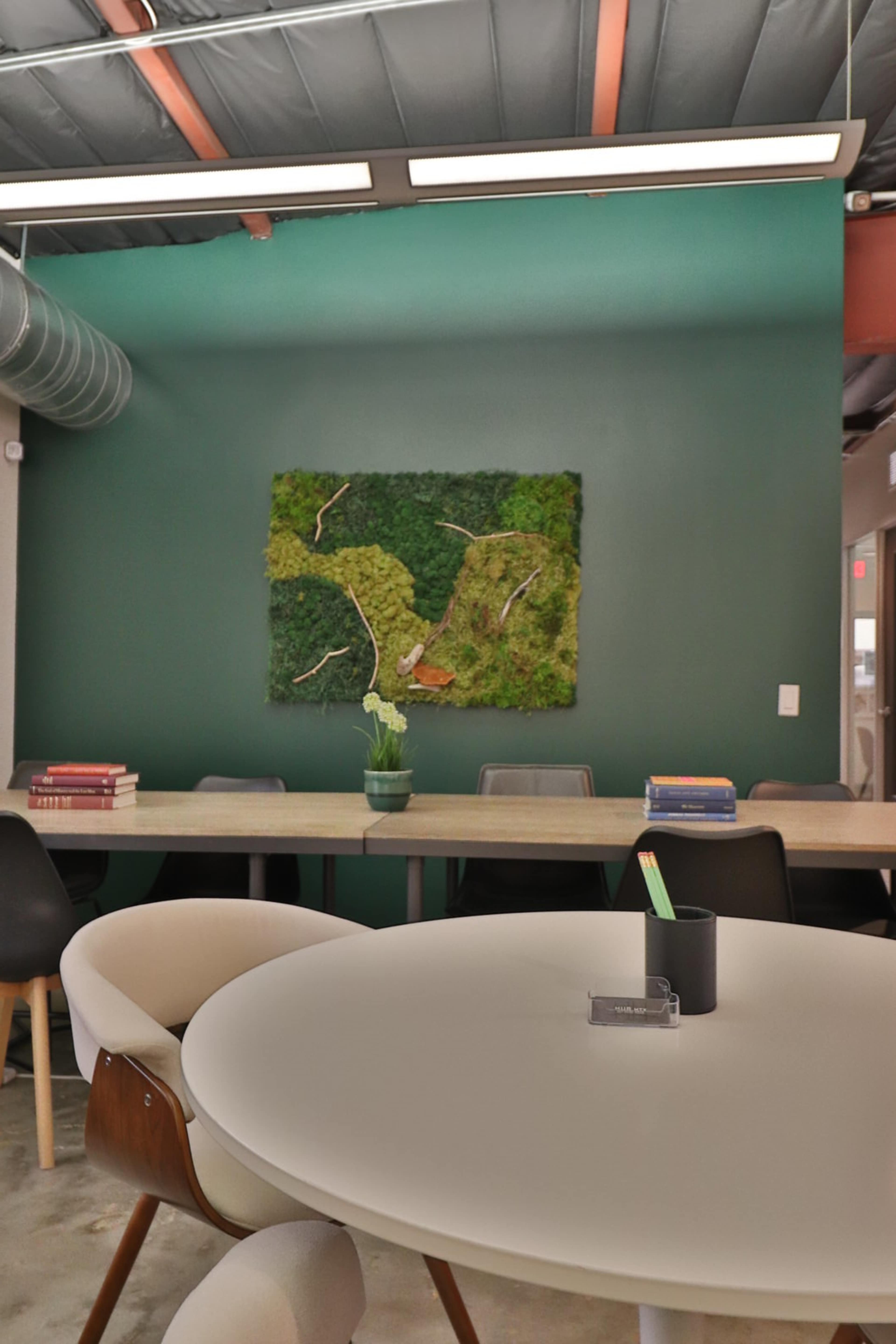 A round white table and chairs are positioned in a modern office space featuring a green accent wall with a textured nature-themed artwork.