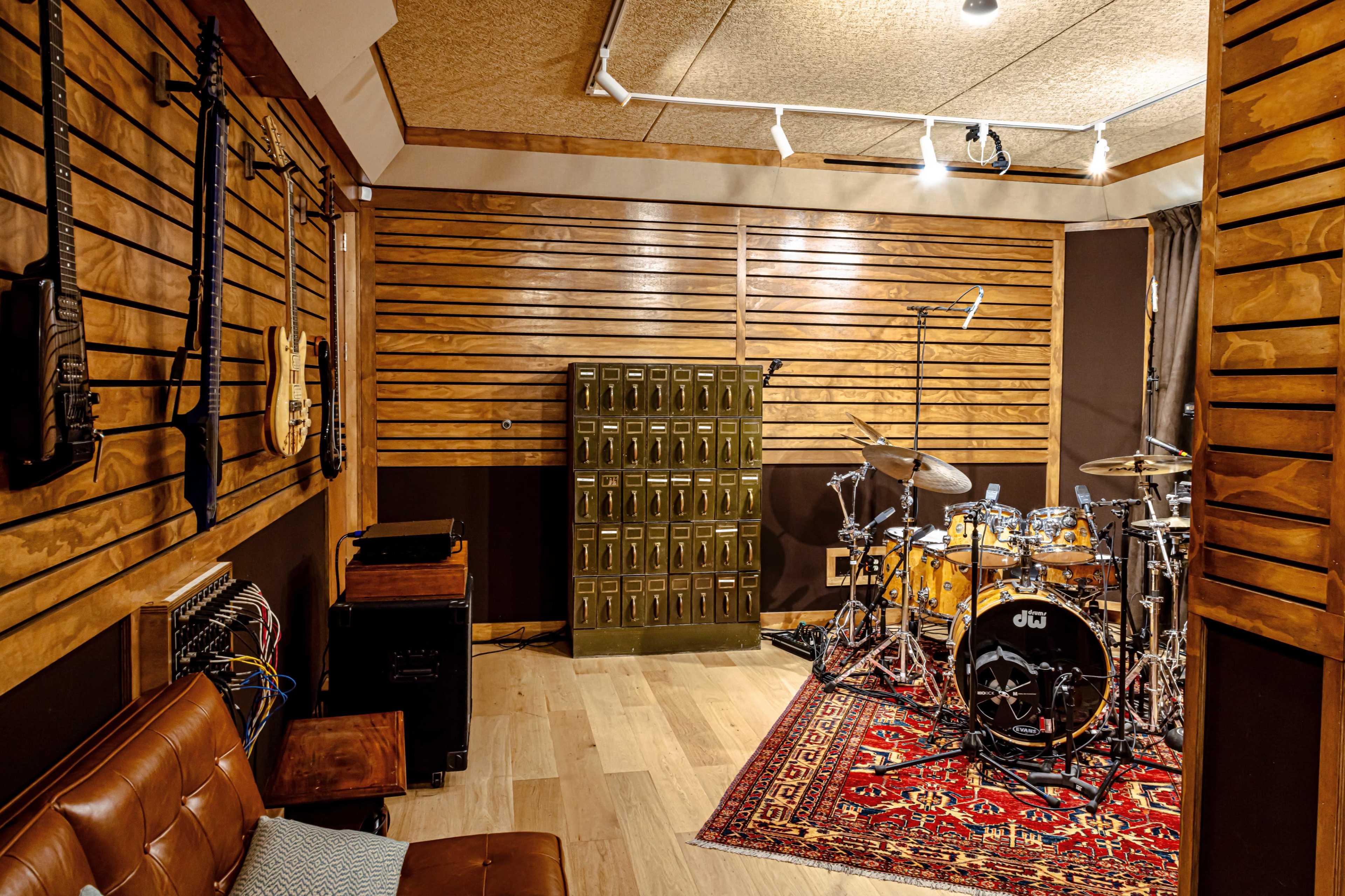 The image shows a recording studio with wooden walls, an acoustic drum set, electric guitars mounted on the wall, and a vintage storage cabinet.