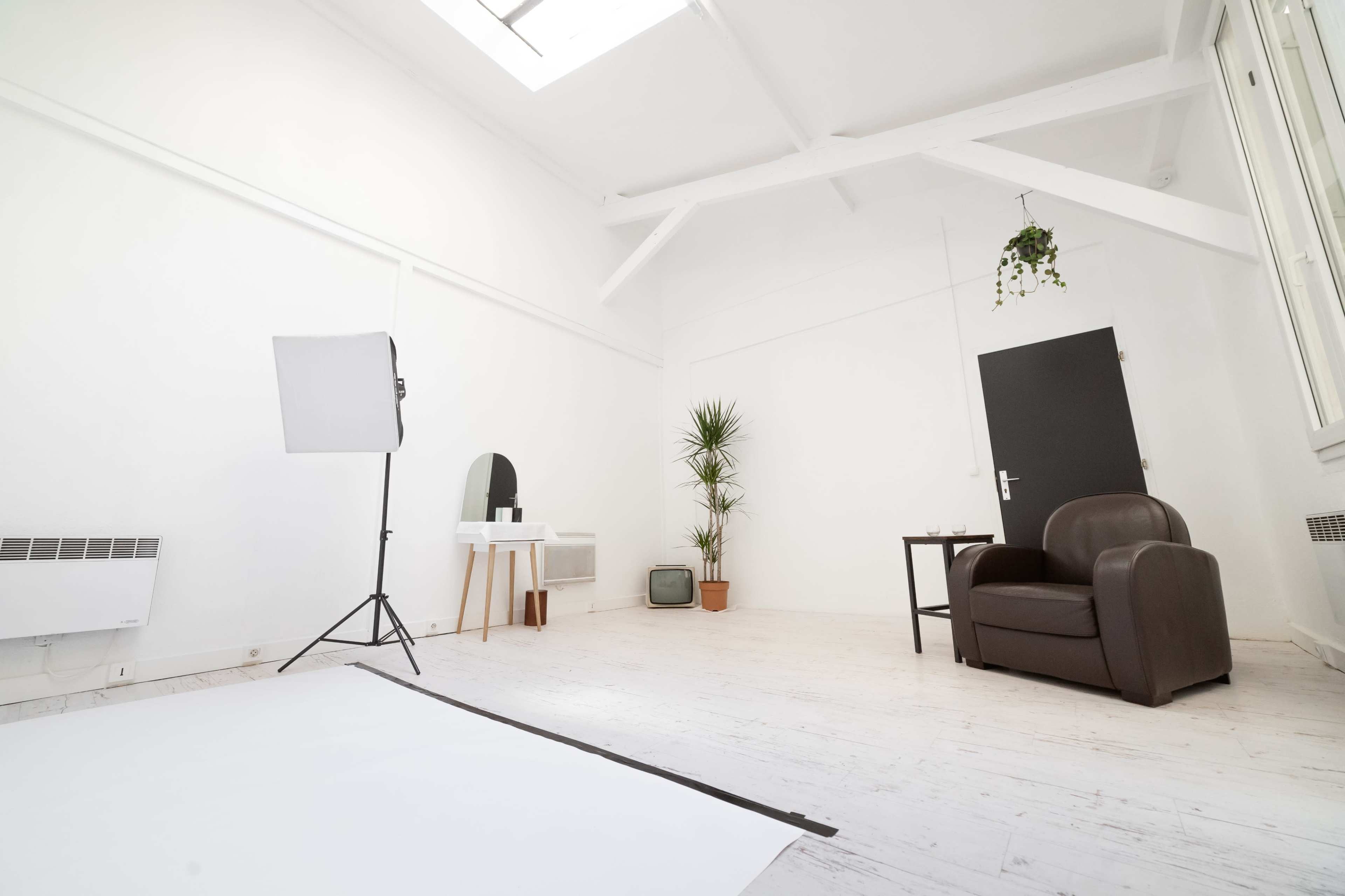 A bright, minimalistic room features a brown armchair, a small table with a mirror, a potted plant, and a softbox light setup against white walls.