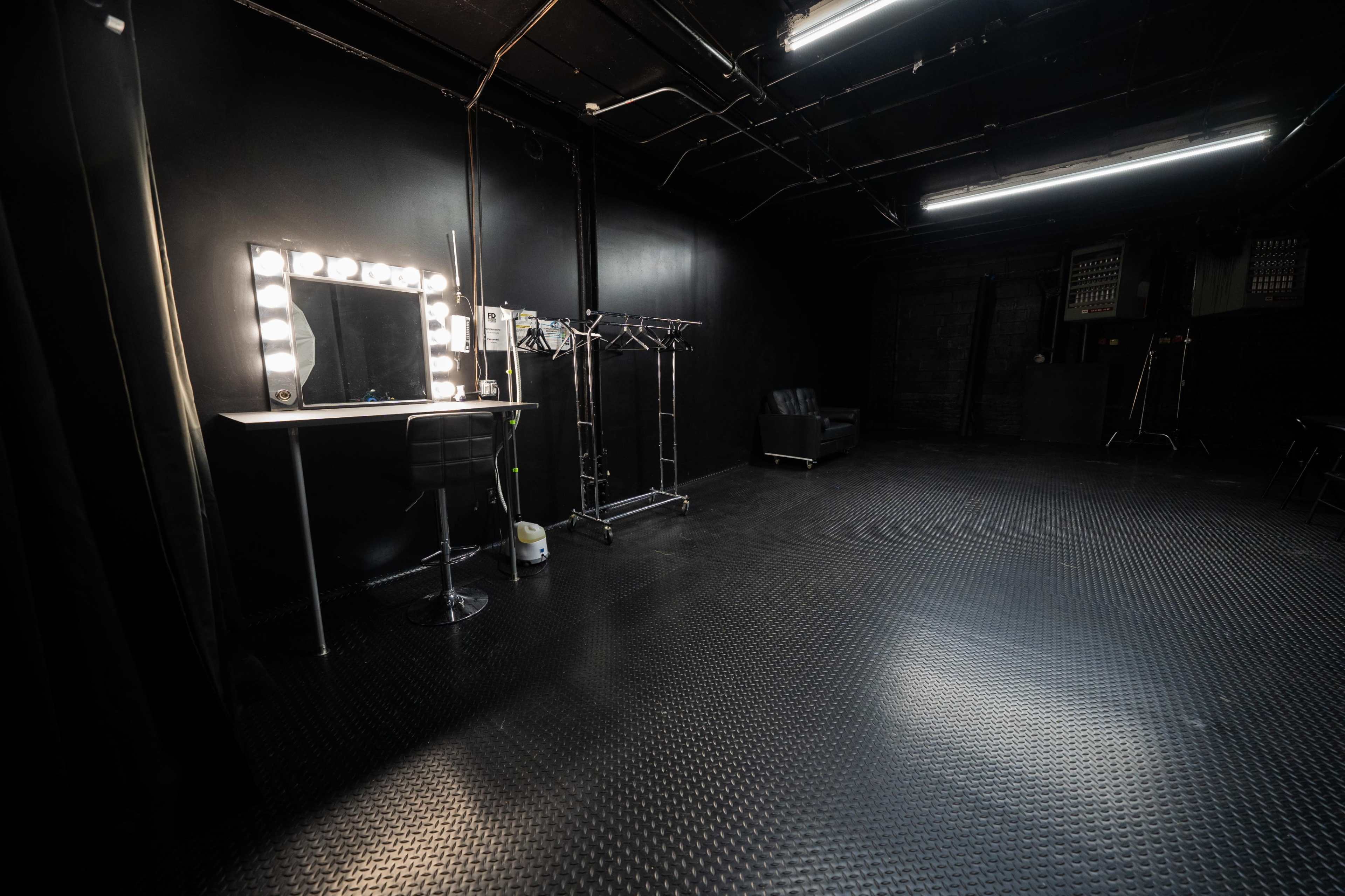 A dimly lit studio space with a makeup mirror, clothing racks, and a black leather chair against a black backdrop.