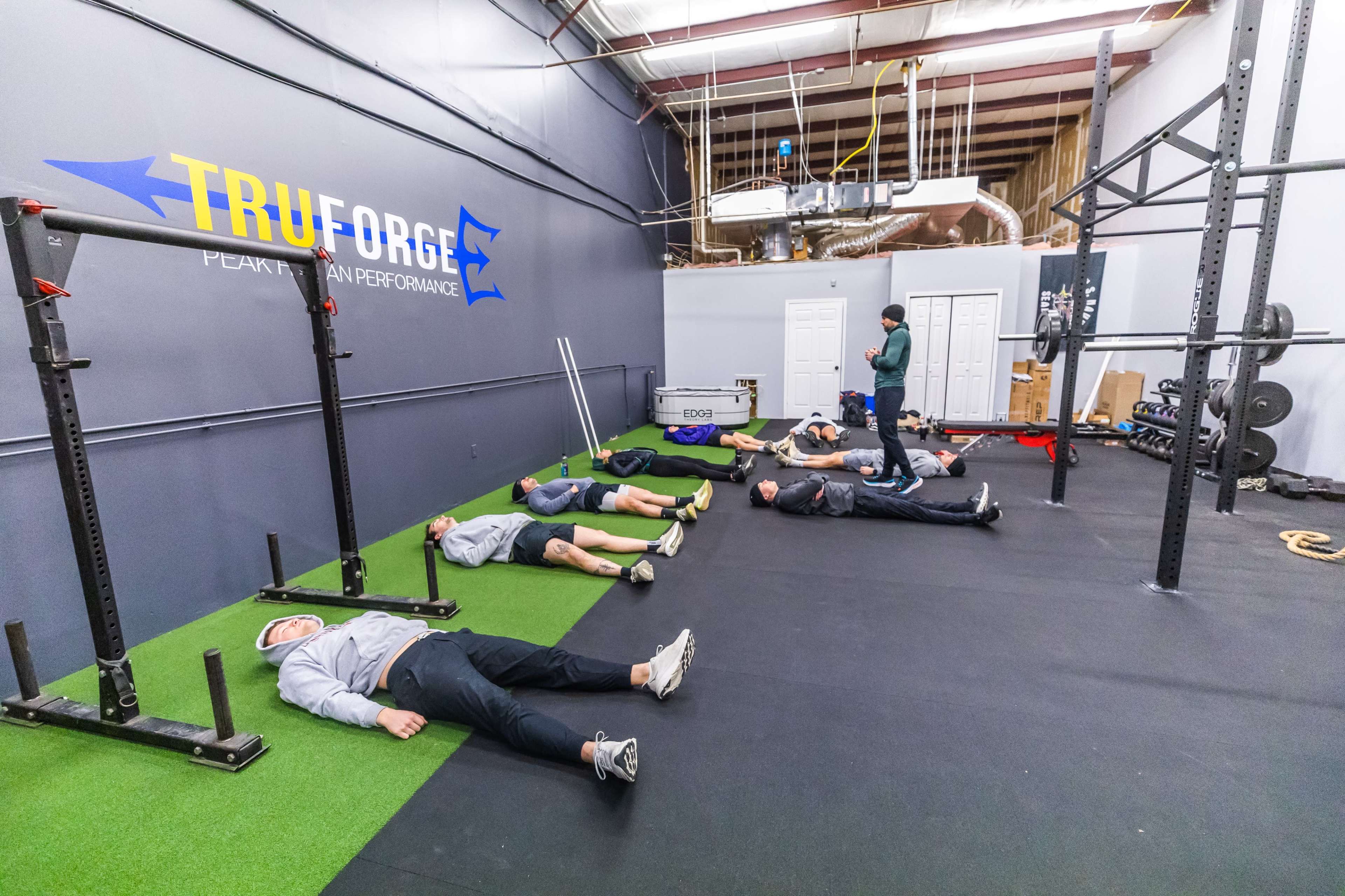 Several people lie on the floor of a gym, while one person stands nearby, in a fitness facility.
