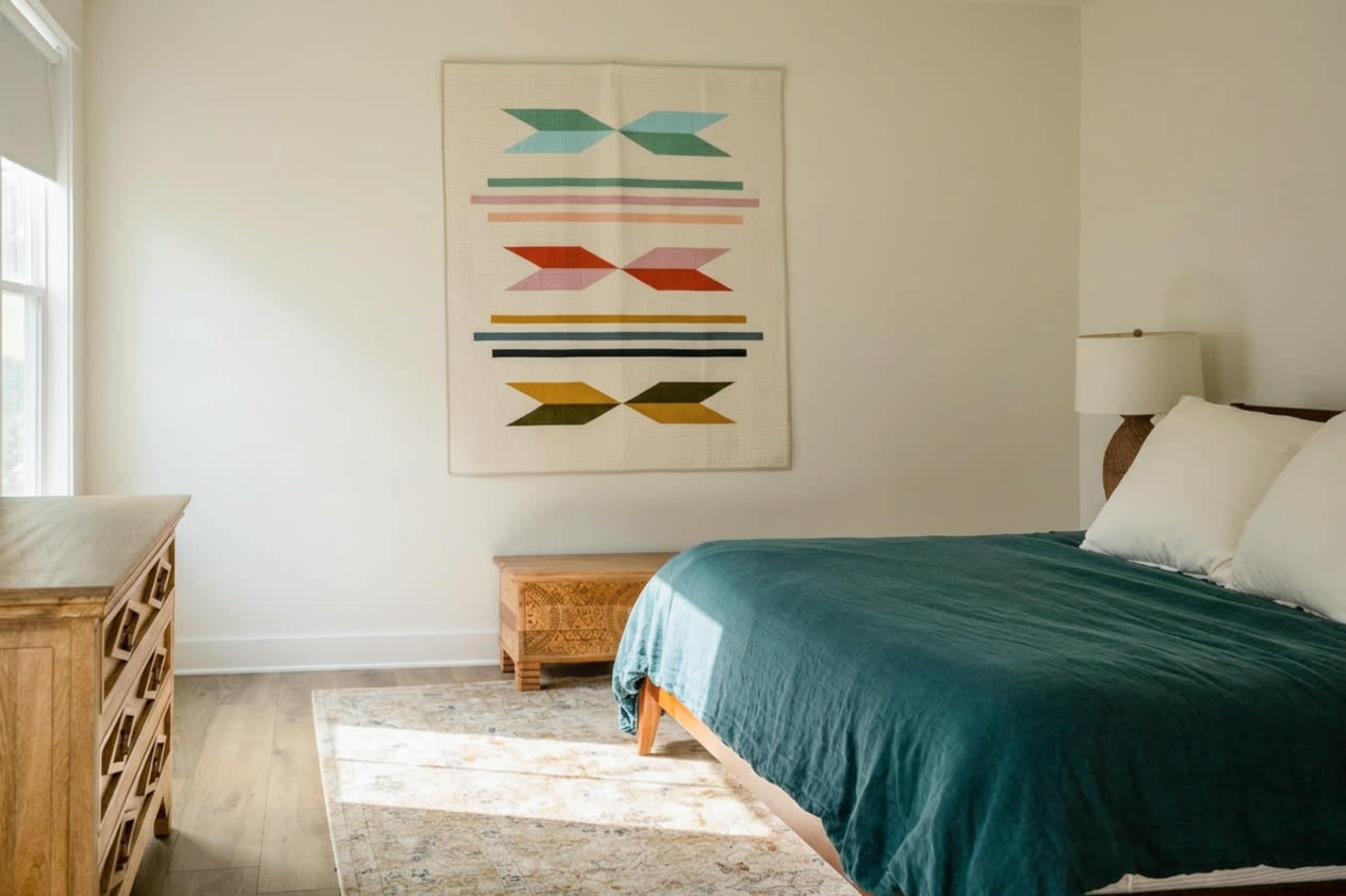 The image shows a neatly arranged bedroom featuring a bed with a teal comforter, a wooden nightstand, a decorative wall hanging, and a light rug on the floor.