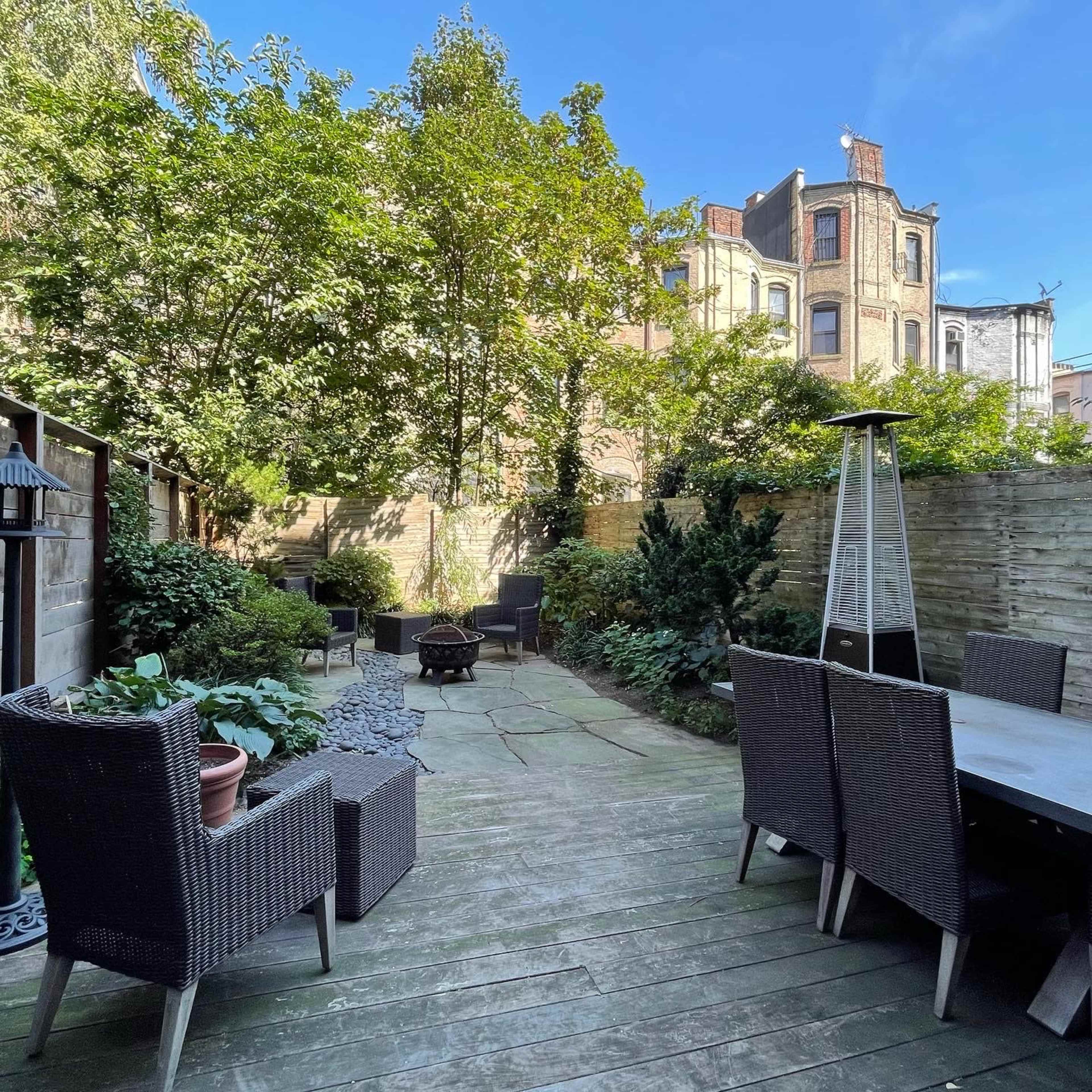 A garden area with stone pavement, surrounded by greenery, featuring outdoor seating and a tall heater.