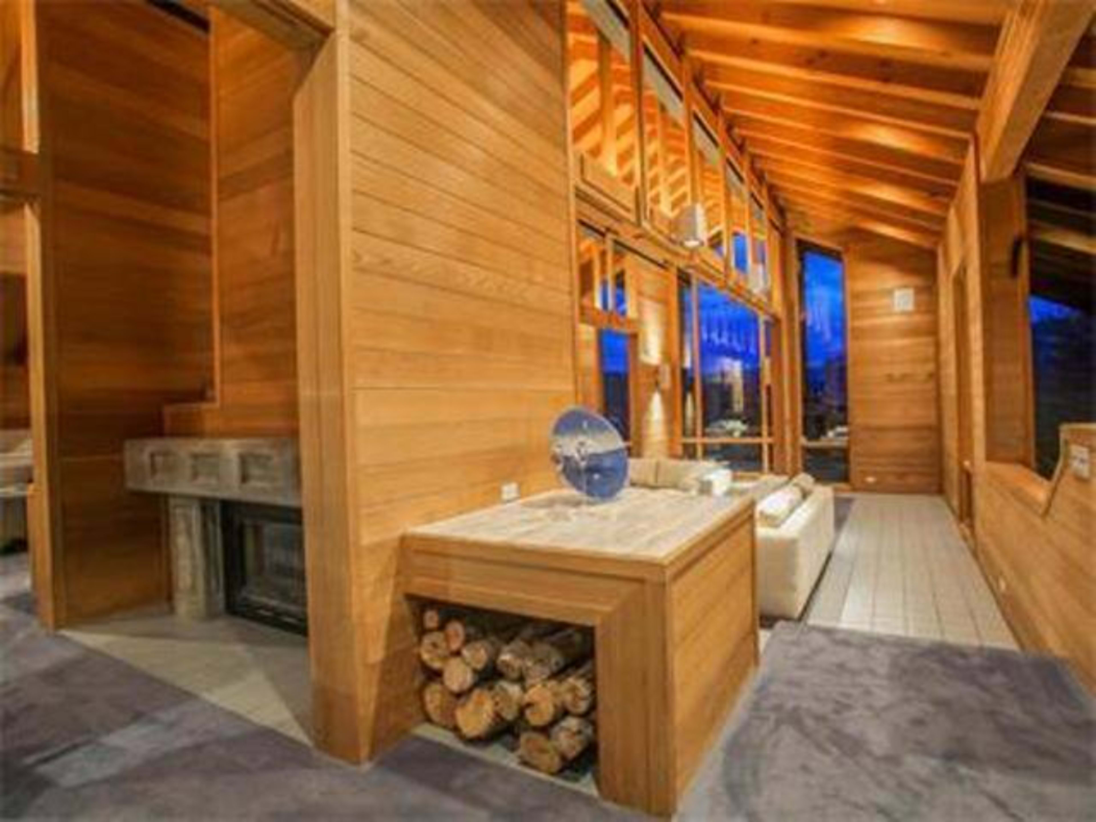The interior of a wooden-paneled living space features large windows, a fireplace, and a stacked log display.