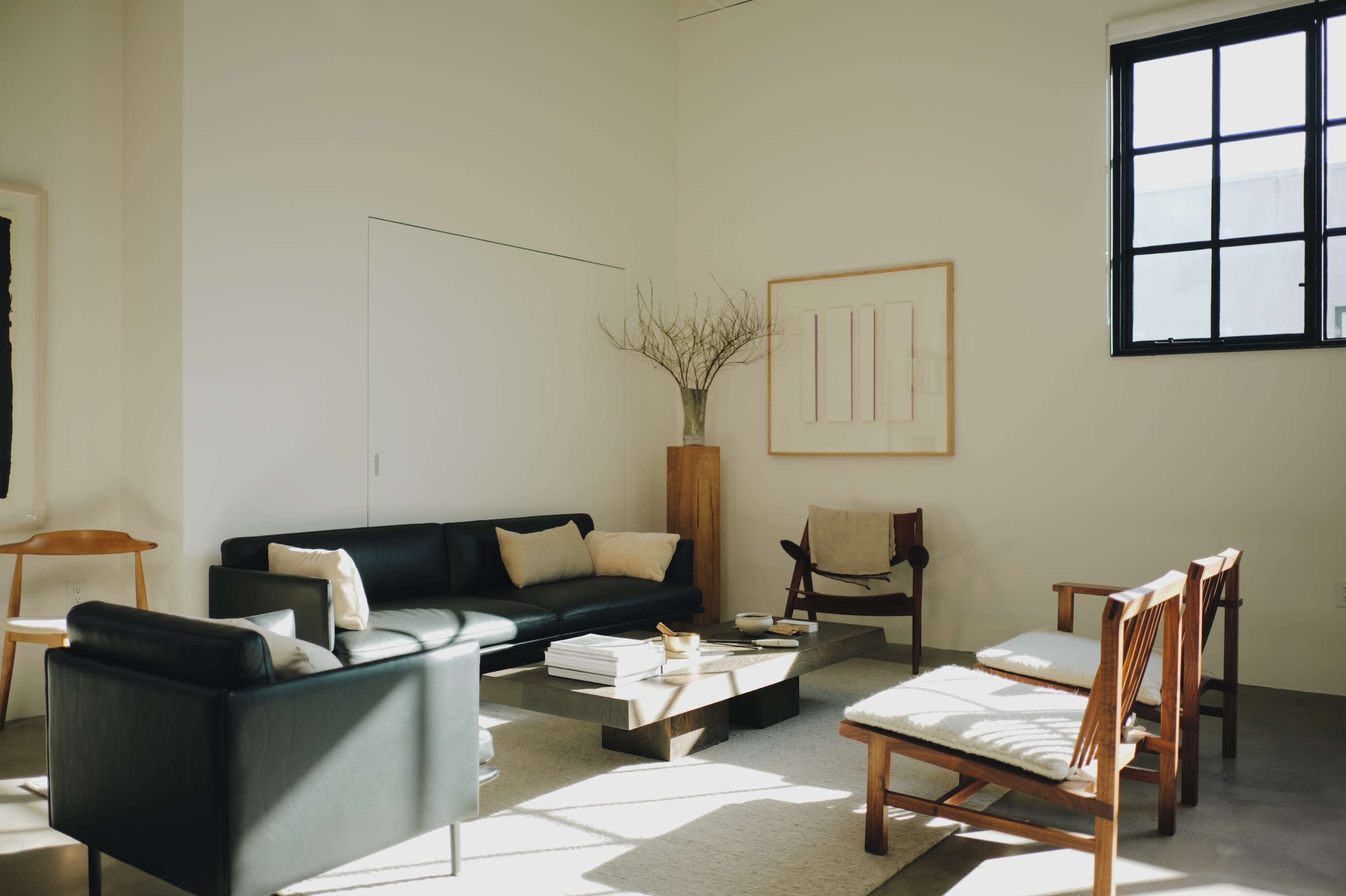 The image shows a modern living room with a black sofa, wooden chairs, and a coffee table on a light-colored rug, illuminated by natural light from a large window.