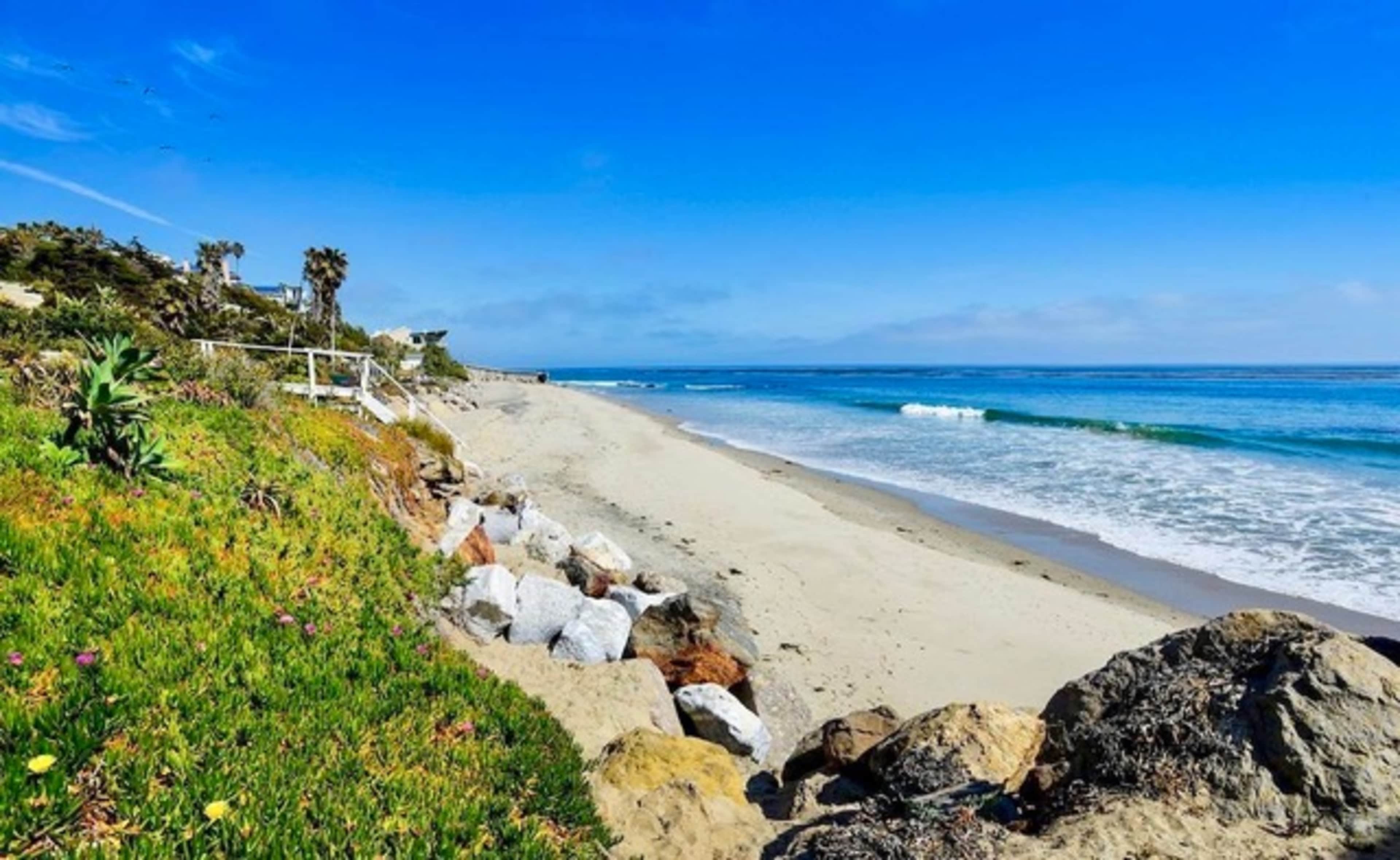 A sandy beach with gentle waves and rocky outcroppings is lined by greenery under a clear blue sky.