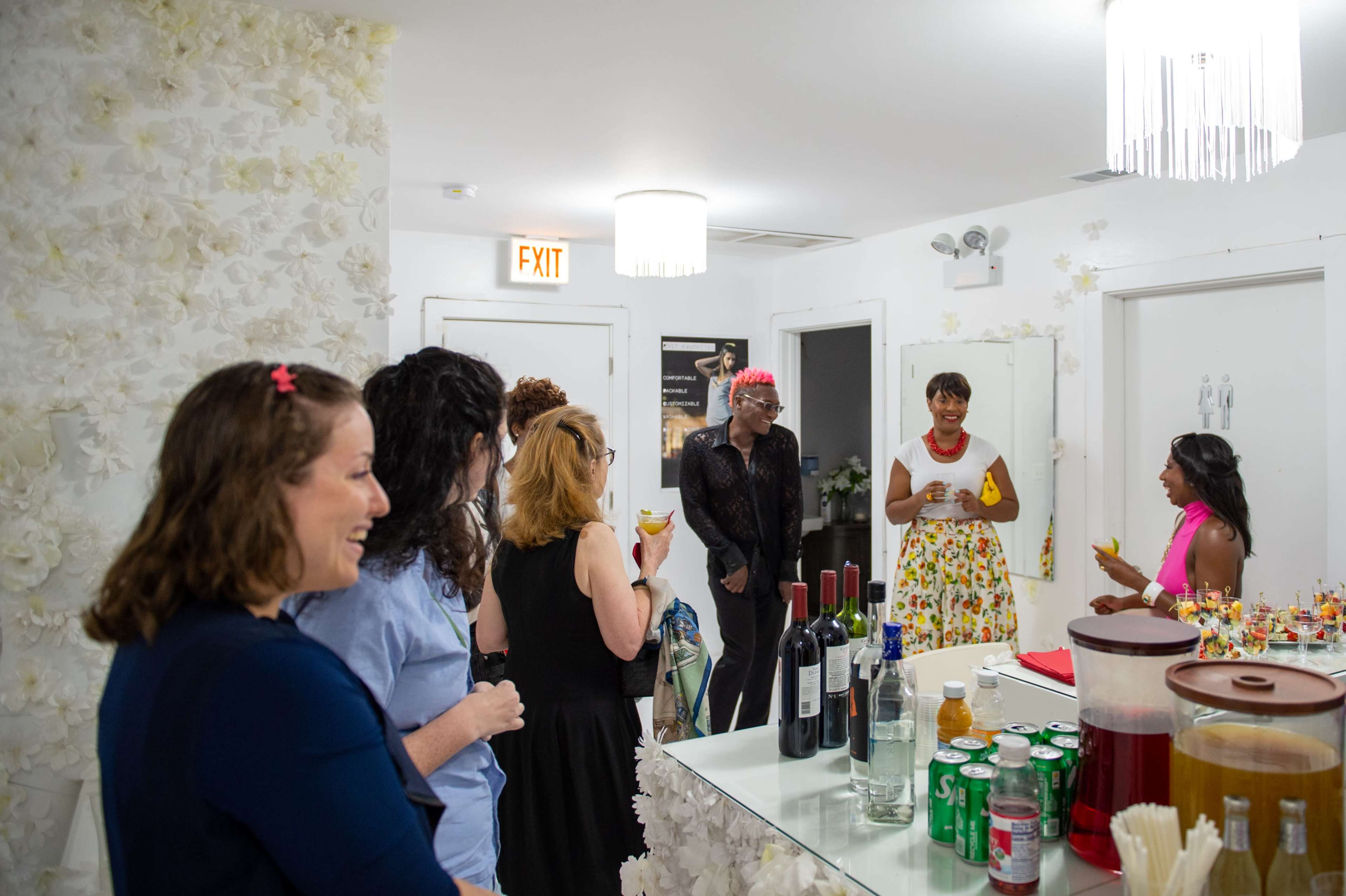 A group of women are gathered in a brightly lit room adorned with decorative flowers, socializing around a bar filled with drinks.