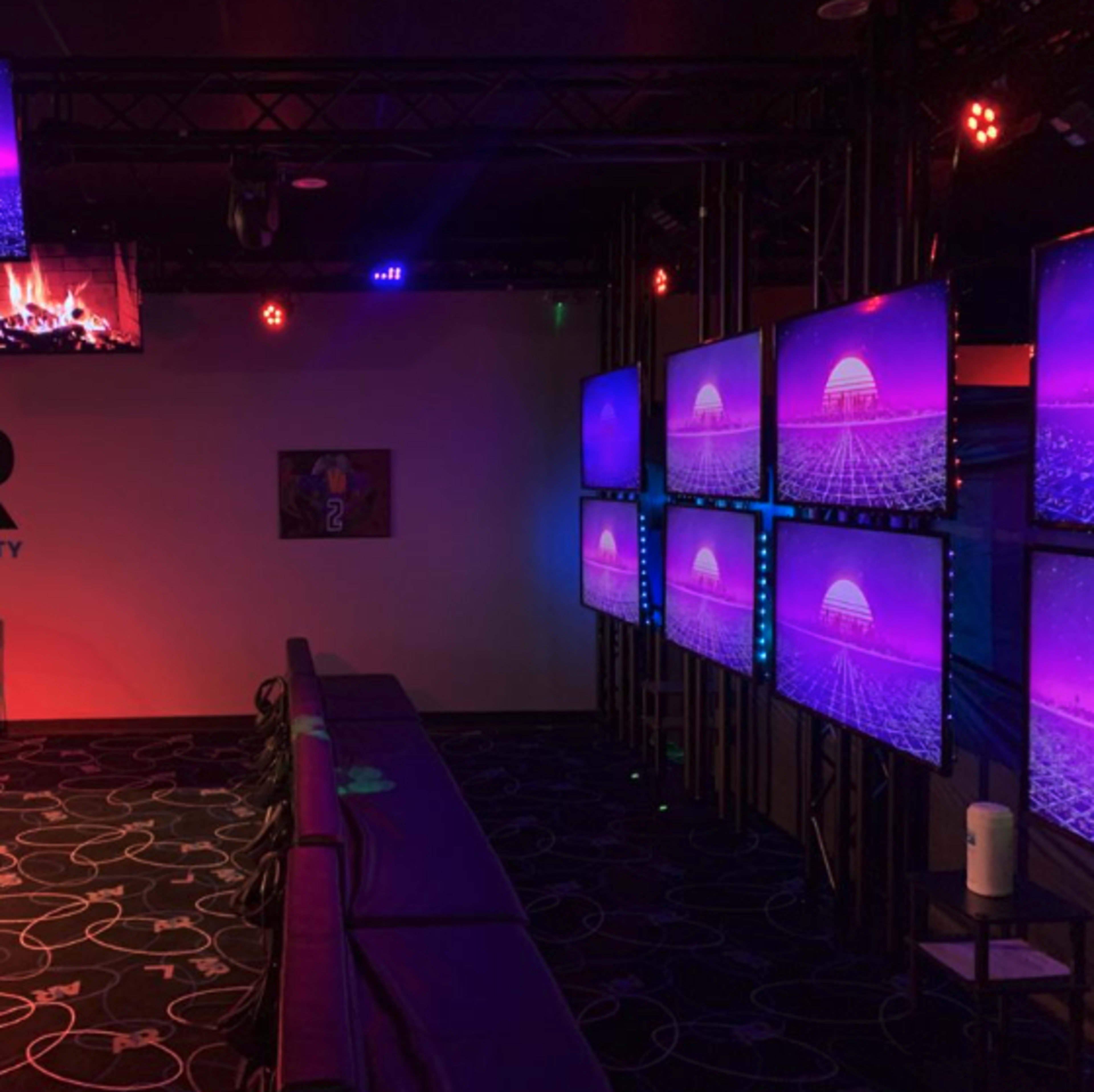 A room with multiple mounted screens displaying a colorful graphic, featuring a digital sunset and geometric patterns, with a row of seating along one side.