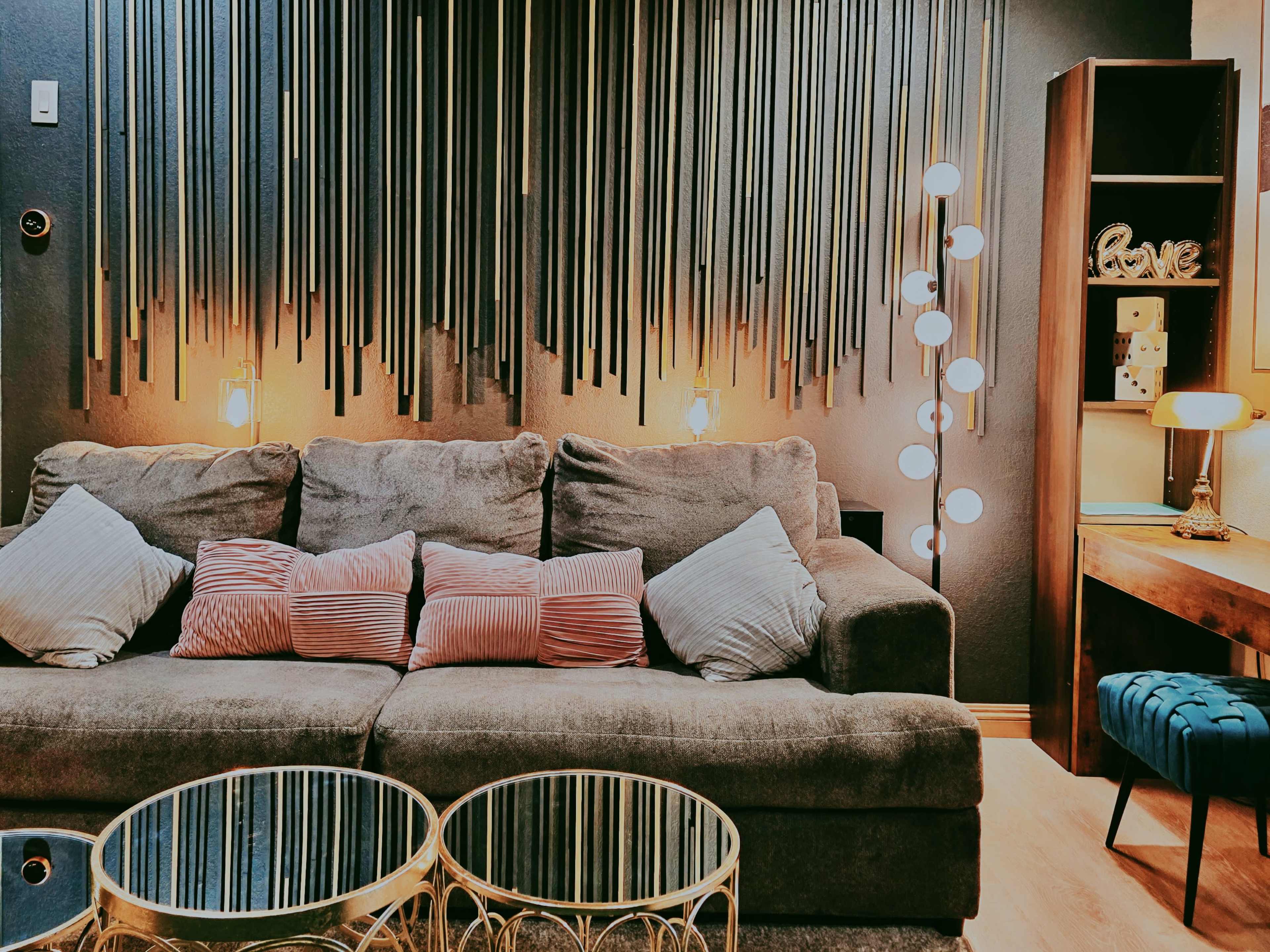 A cozy living room features a gray sofa with decorative pillows, a pair of gold-framed coffee tables, and warm lighting from floor and wall lamps.
