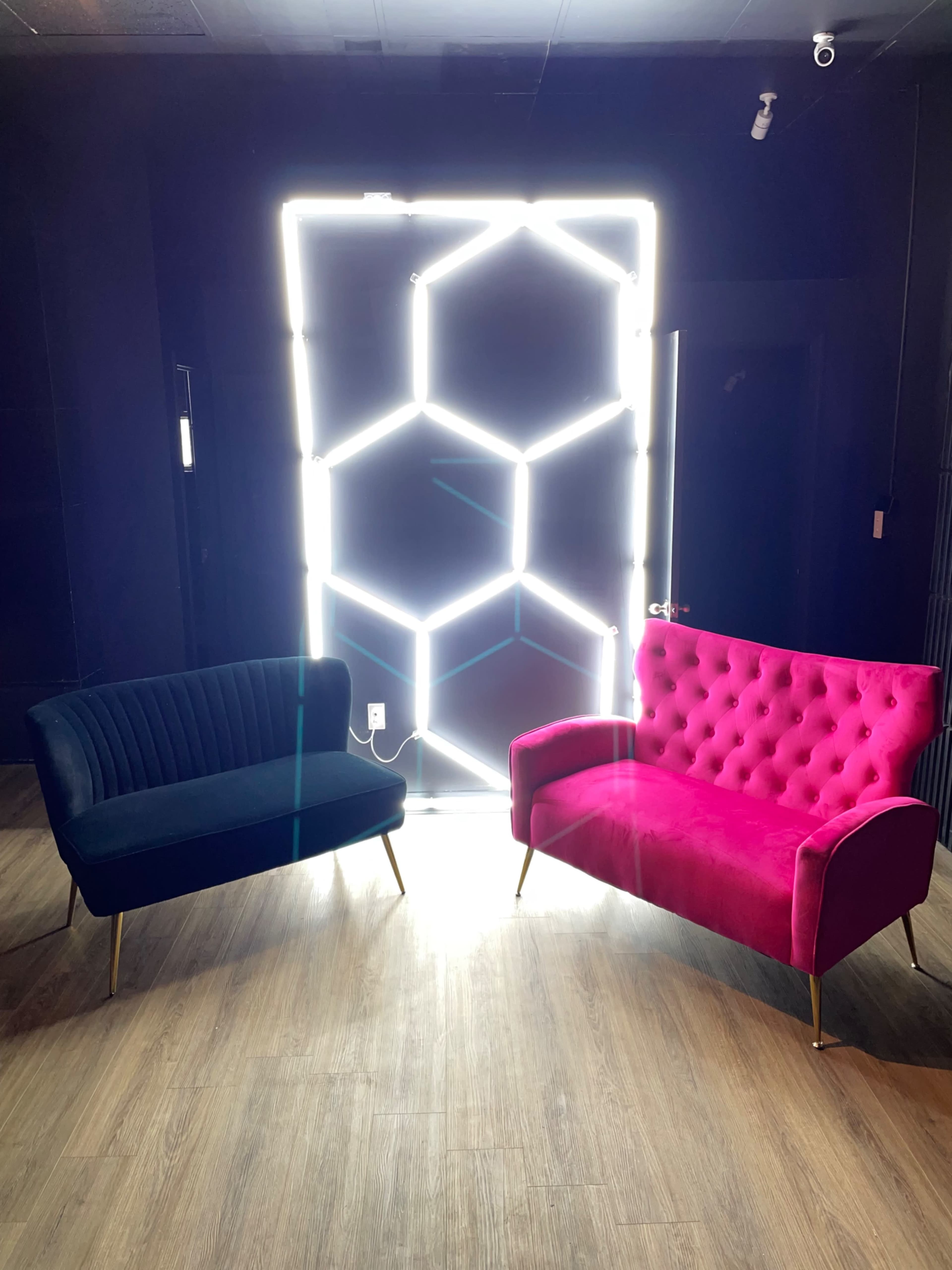 Two stylish couches, one black with vertical stripes and one pink with tufted upholstery, positioned in front of a hexagonal neon light installation against a dark background.