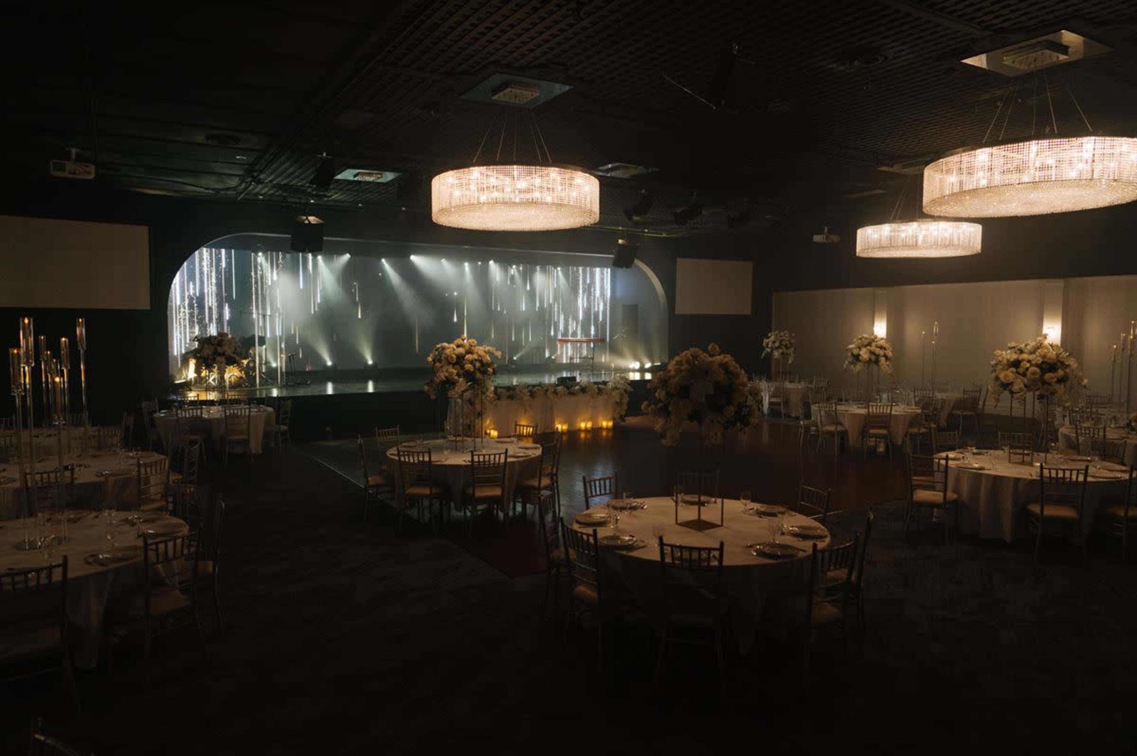 The image shows an elegant indoor event space featuring round dining tables with white tablecloths, floral centerpieces, and a stage illuminated with a backdrop of soft lights.