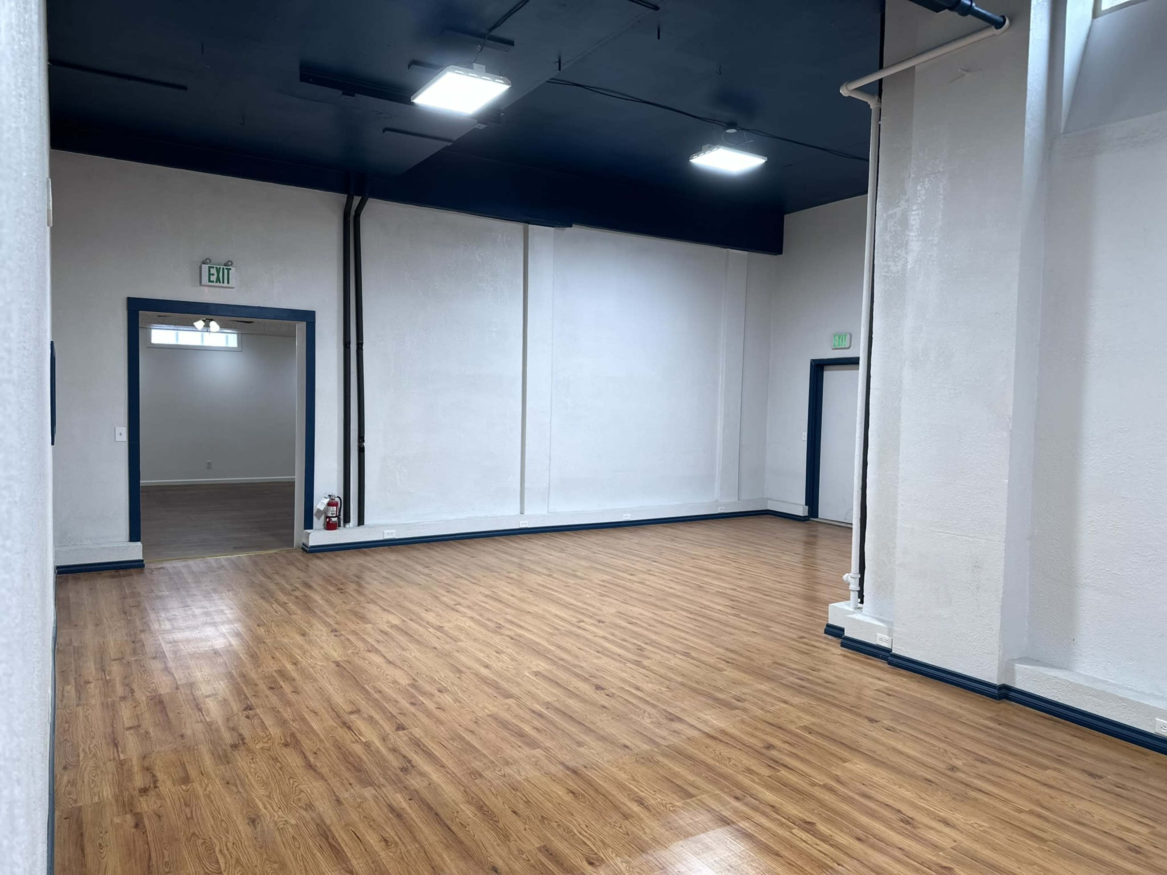 A spacious, empty room with wooden flooring, white walls, and black ceiling tiles, featuring a doorway leading to another area.