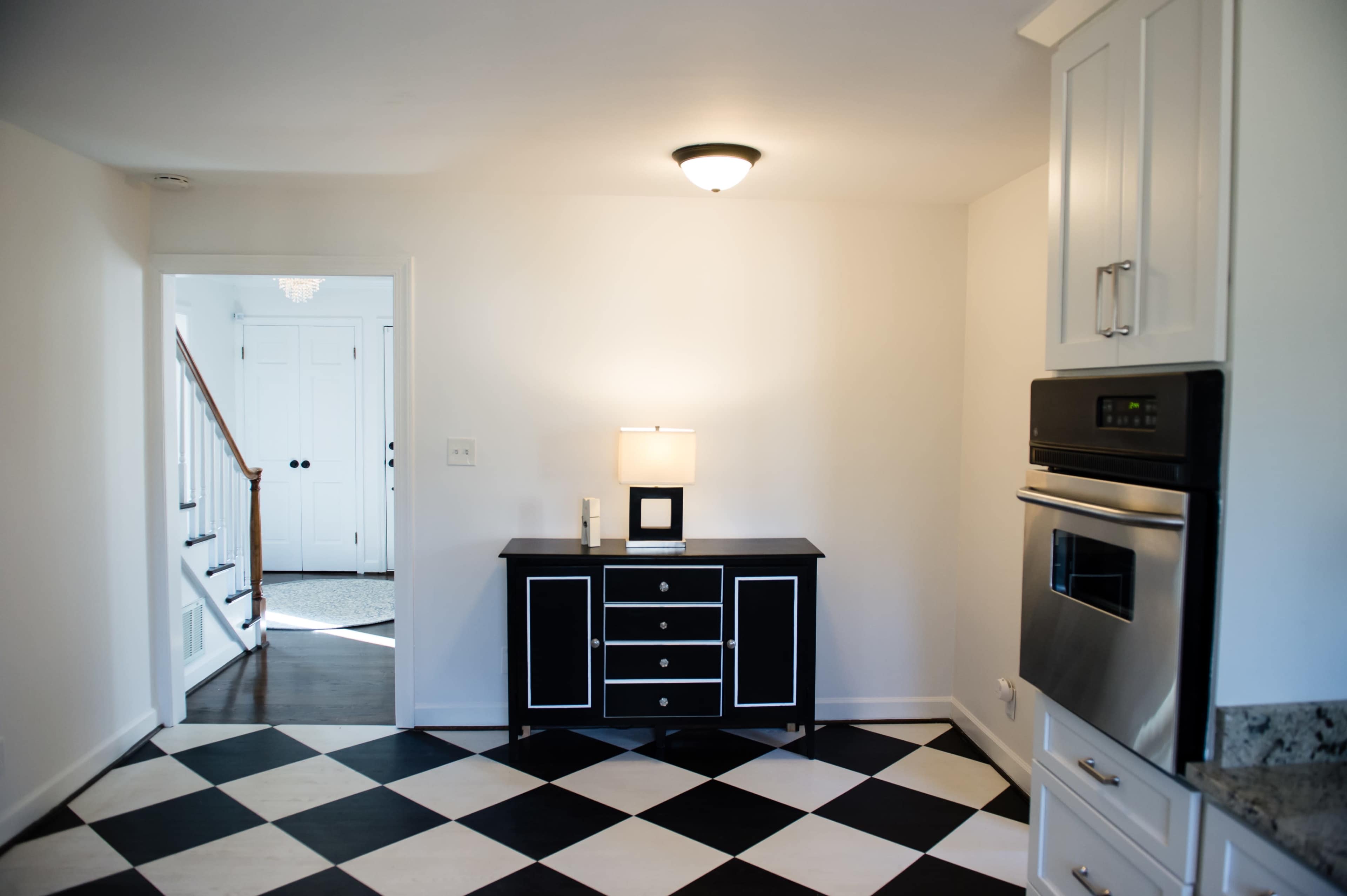 A modern kitchen features black and white checkered flooring, a dark cabinet with a lamp, and an entryway leading to a staircase.