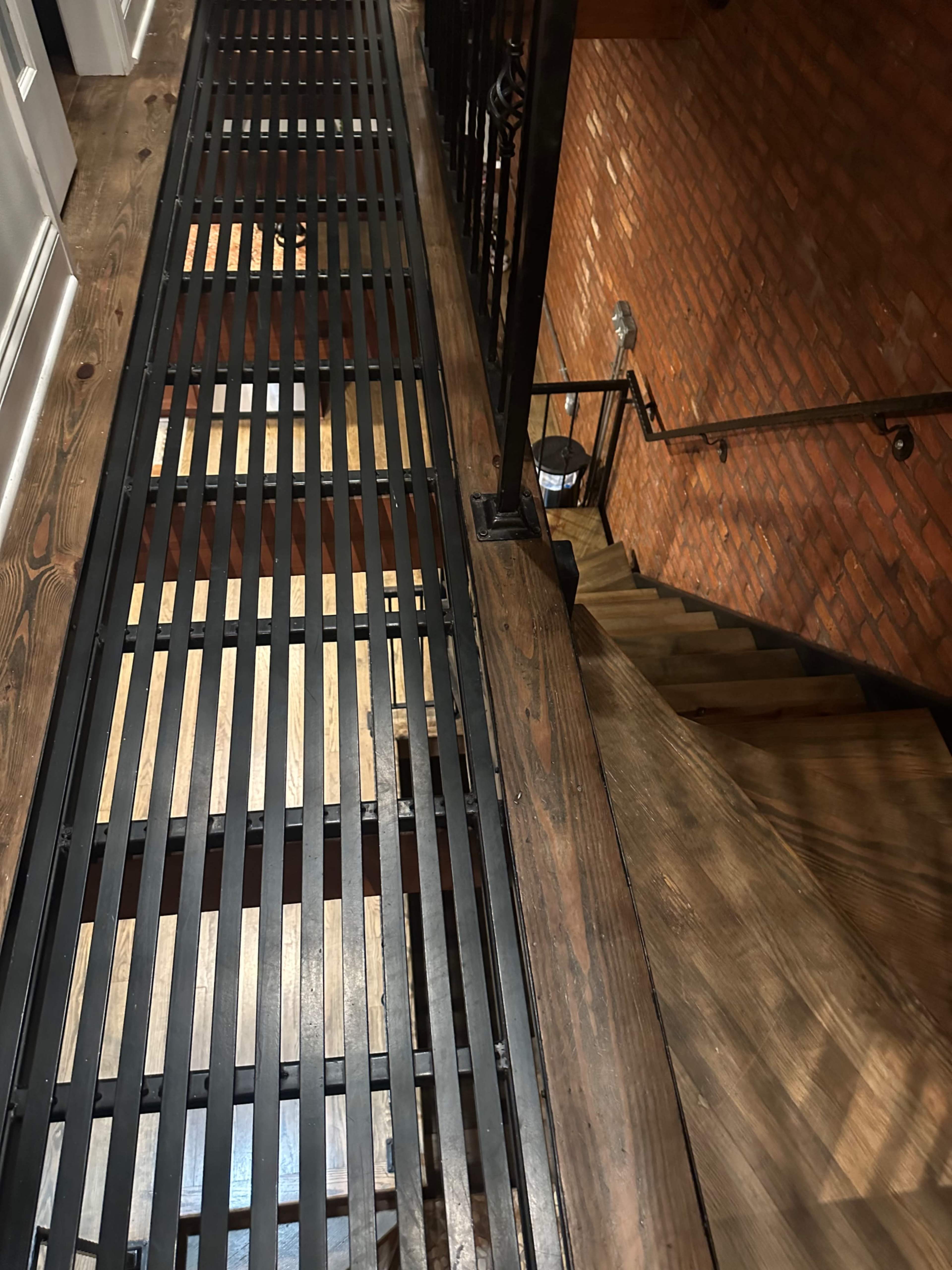 The image shows a metal grate walkway overlooking a staircase with wooden steps and a brick wall.