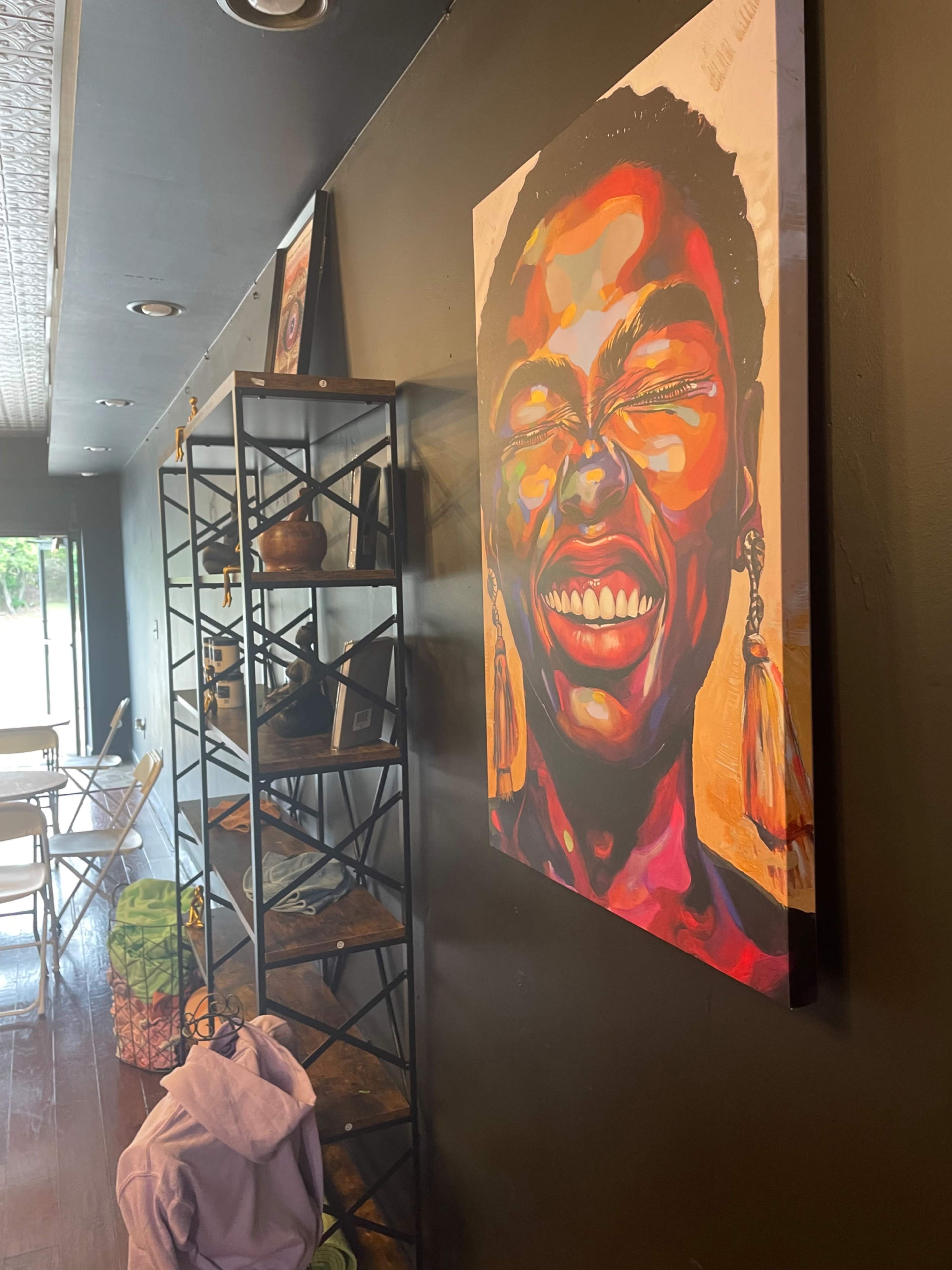 The image shows a brightly colored portrait of a woman smiling on a dark wall, with shelves displaying various items and a seating area in the background.