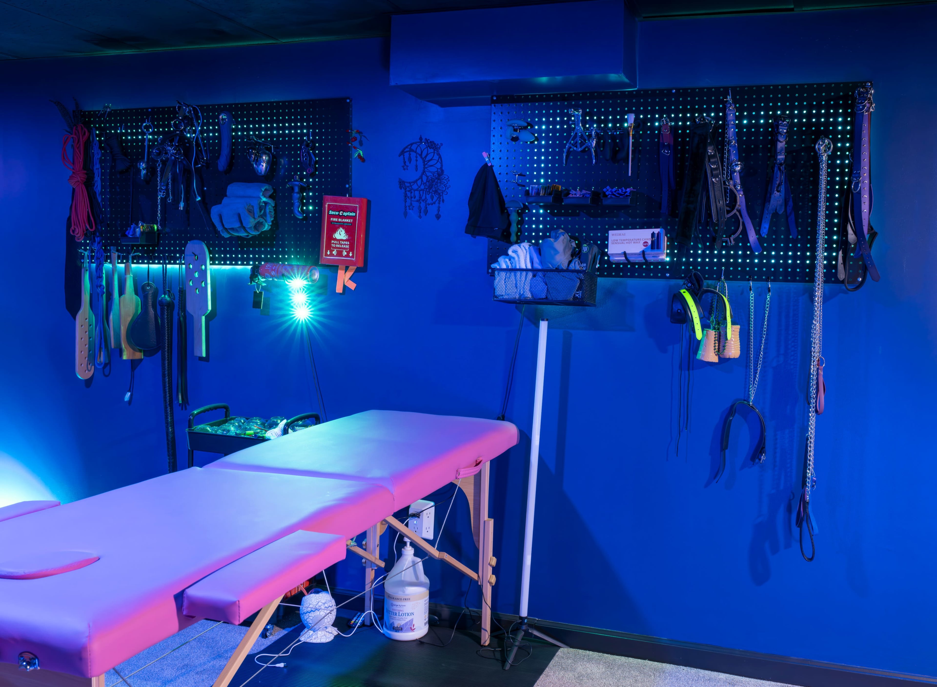 The image shows a massage table in a room with blue walls and various tools and equipment hanging on pegboards behind it.