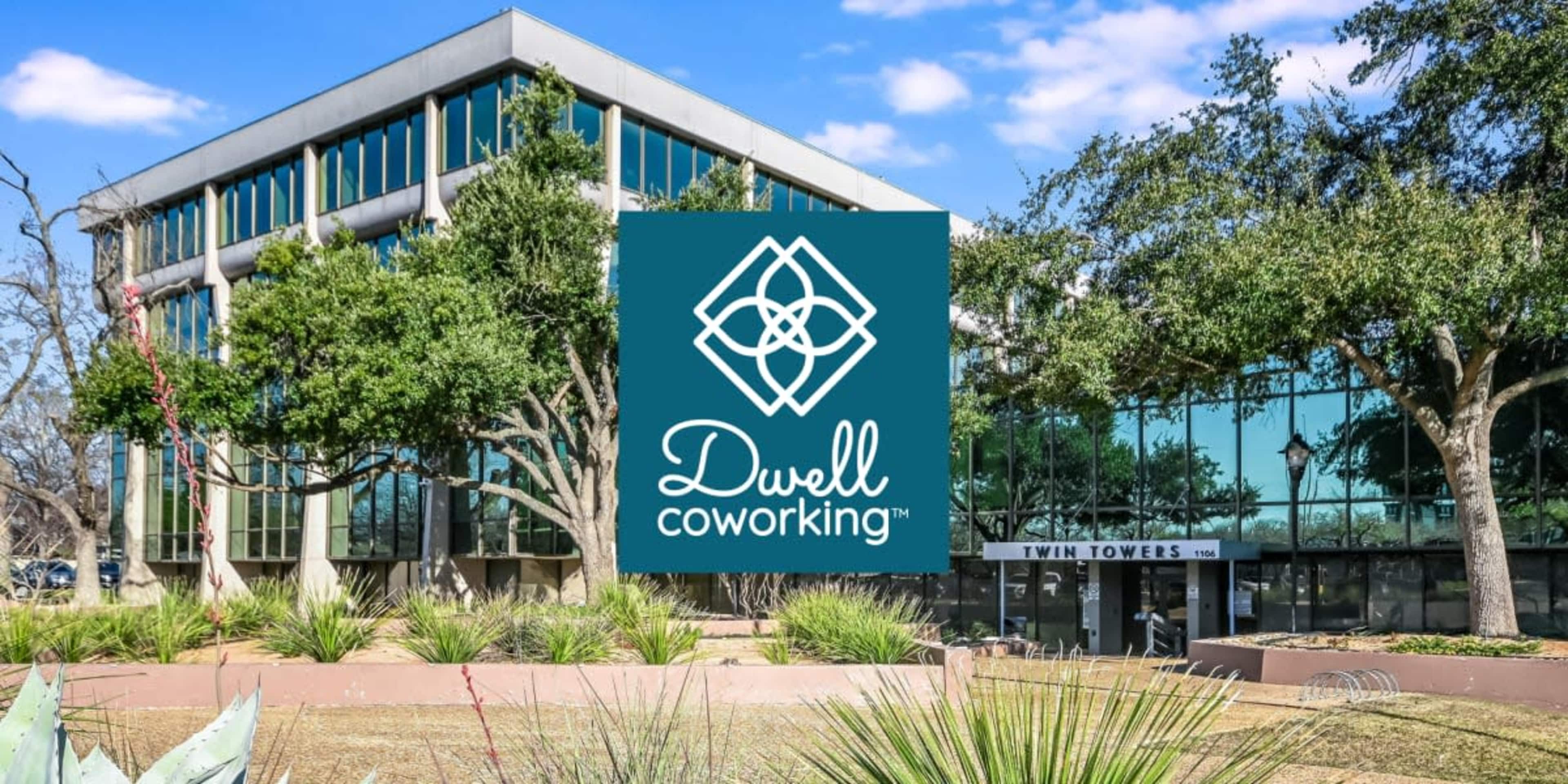 The image shows a modern glass building with the name "Dwell Coworking" prominently displayed in the foreground, surrounded by landscaped greenery.