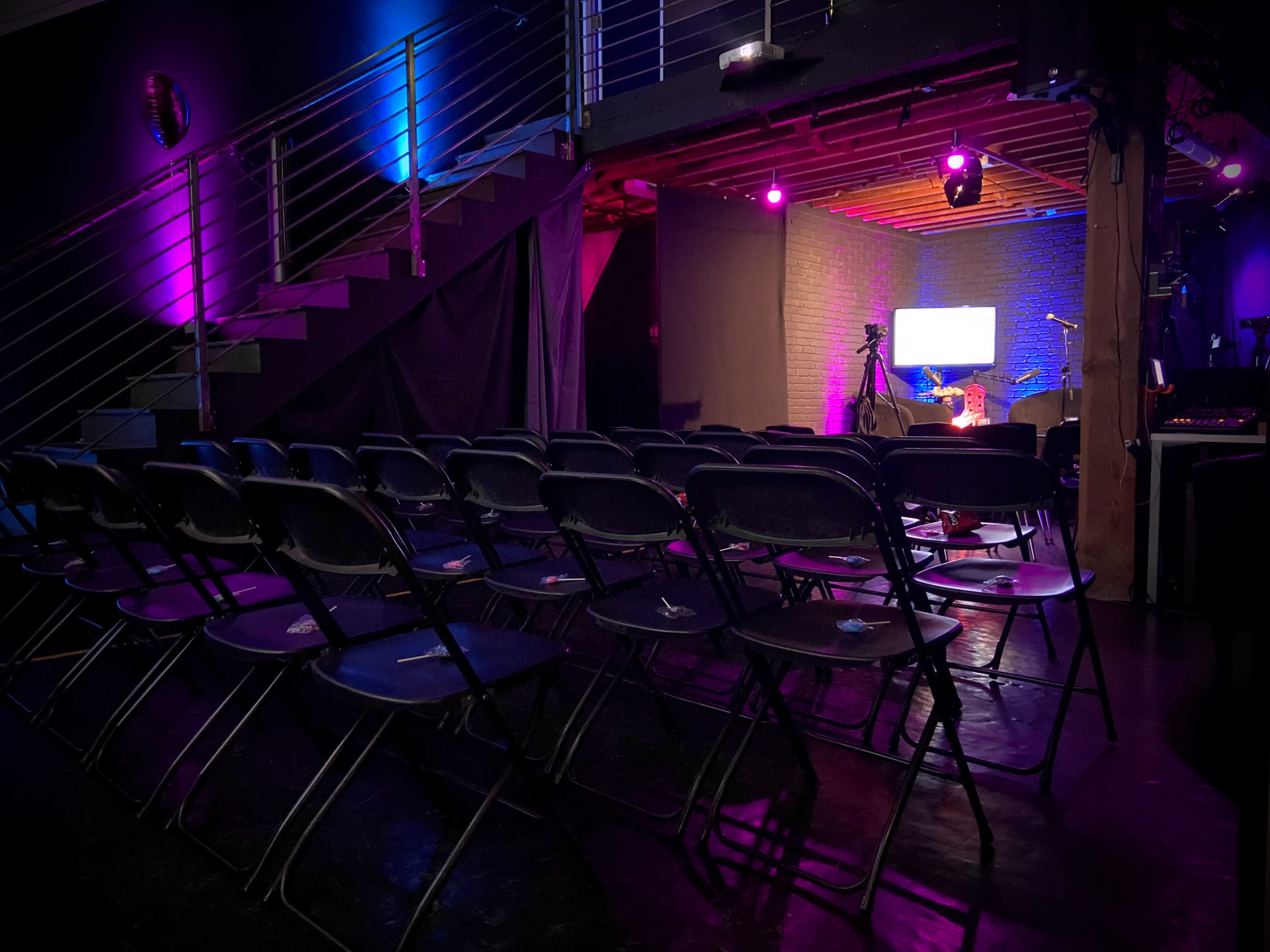 A dimly lit performance space with rows of empty black chairs facing a small stage equipped with a microphone and instruments, framed by purple stage lighting.