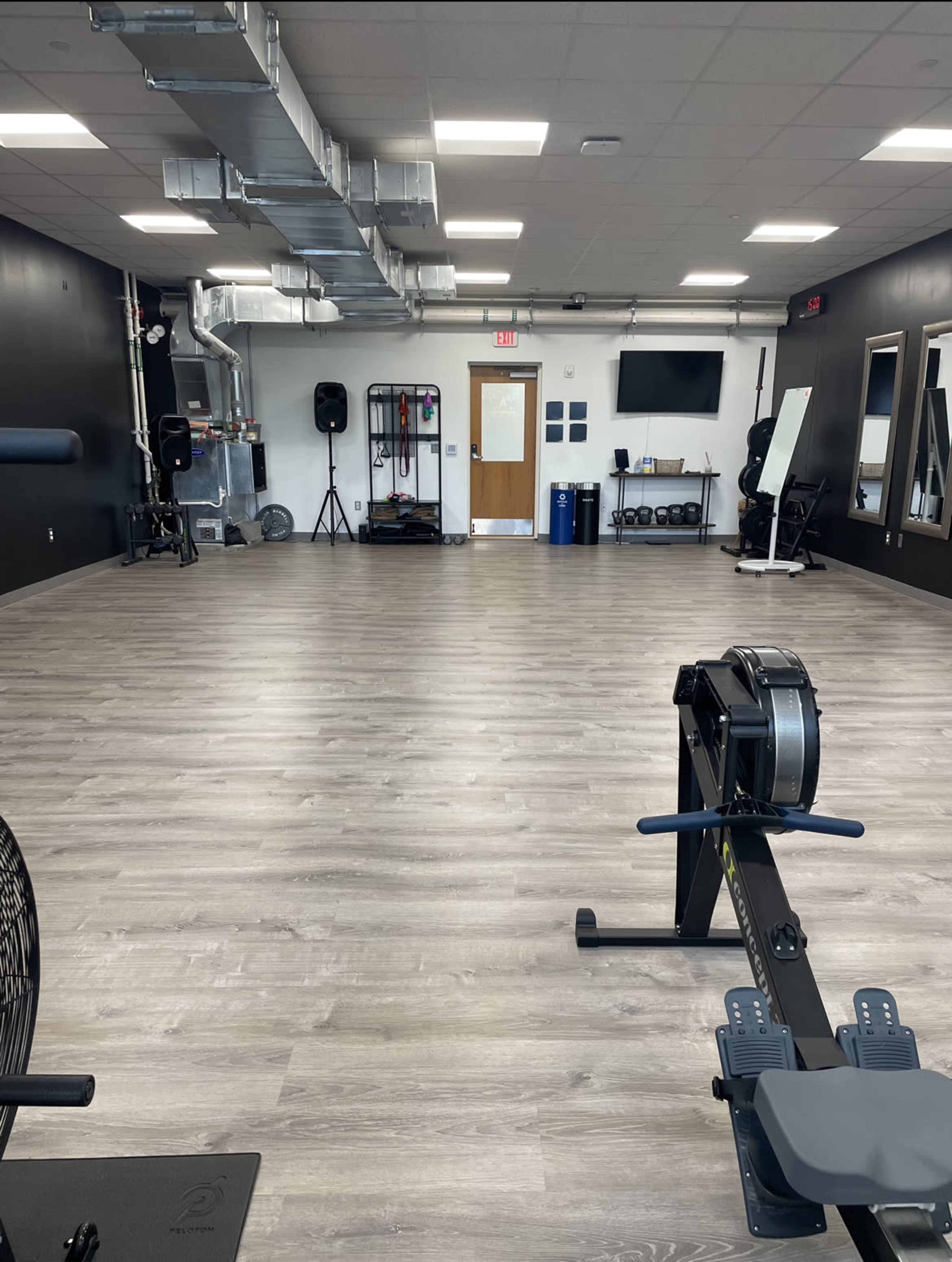 A spacious workout room equipped with exercise machines, mirrors, and various fitness equipment against black walls and wooden flooring.