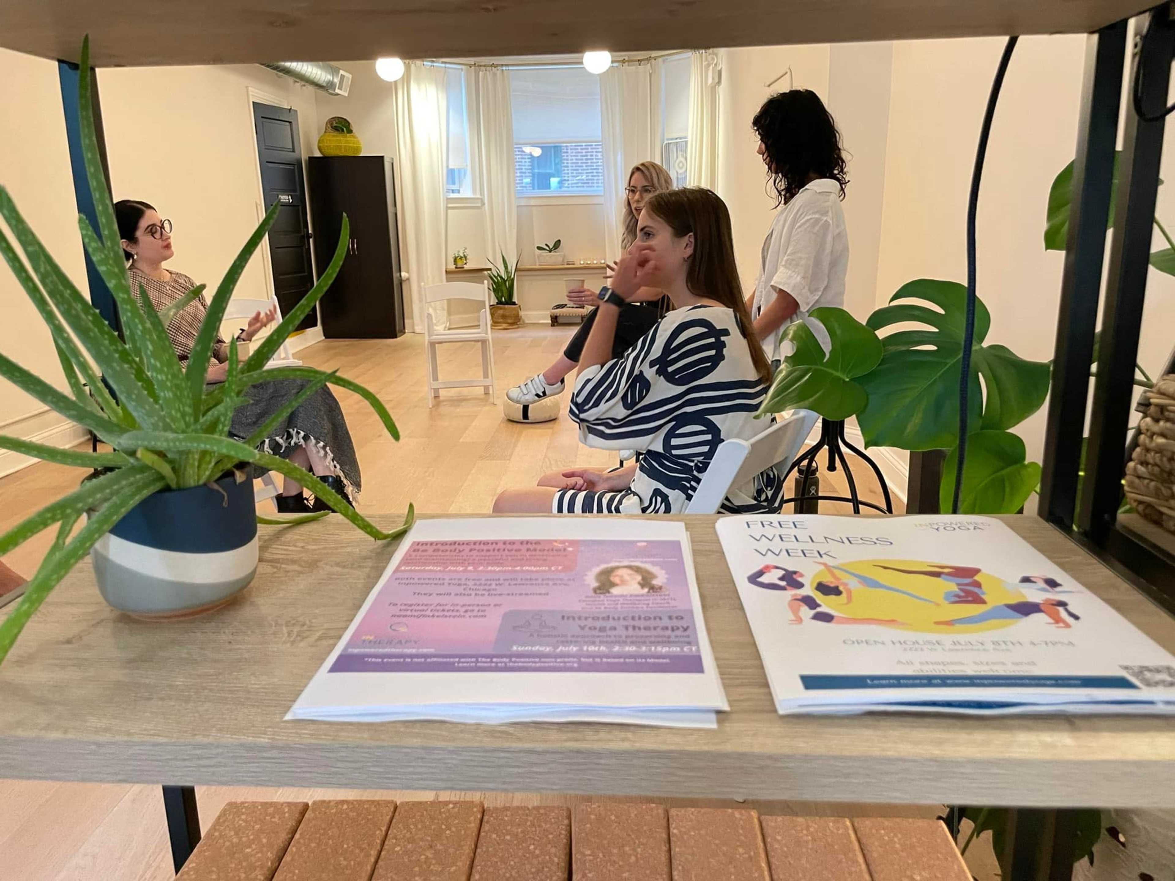 A group of three people sits on chairs in a well-lit room with plants and promotional materials on a shelf in the foreground.