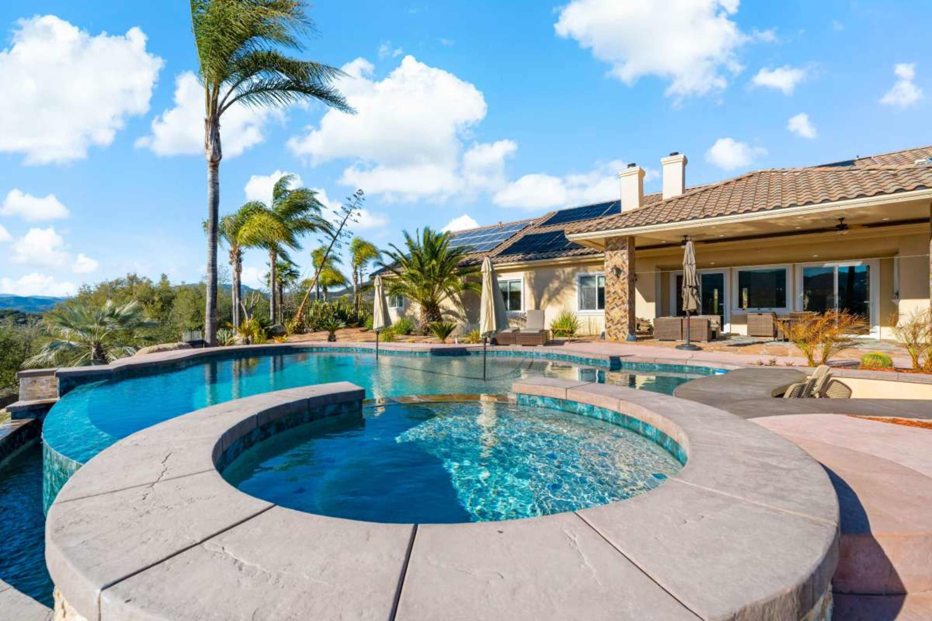 The image features a luxurious backyard pool area with a spa, surrounded by palm trees and a modern home in the background against a clear blue sky.