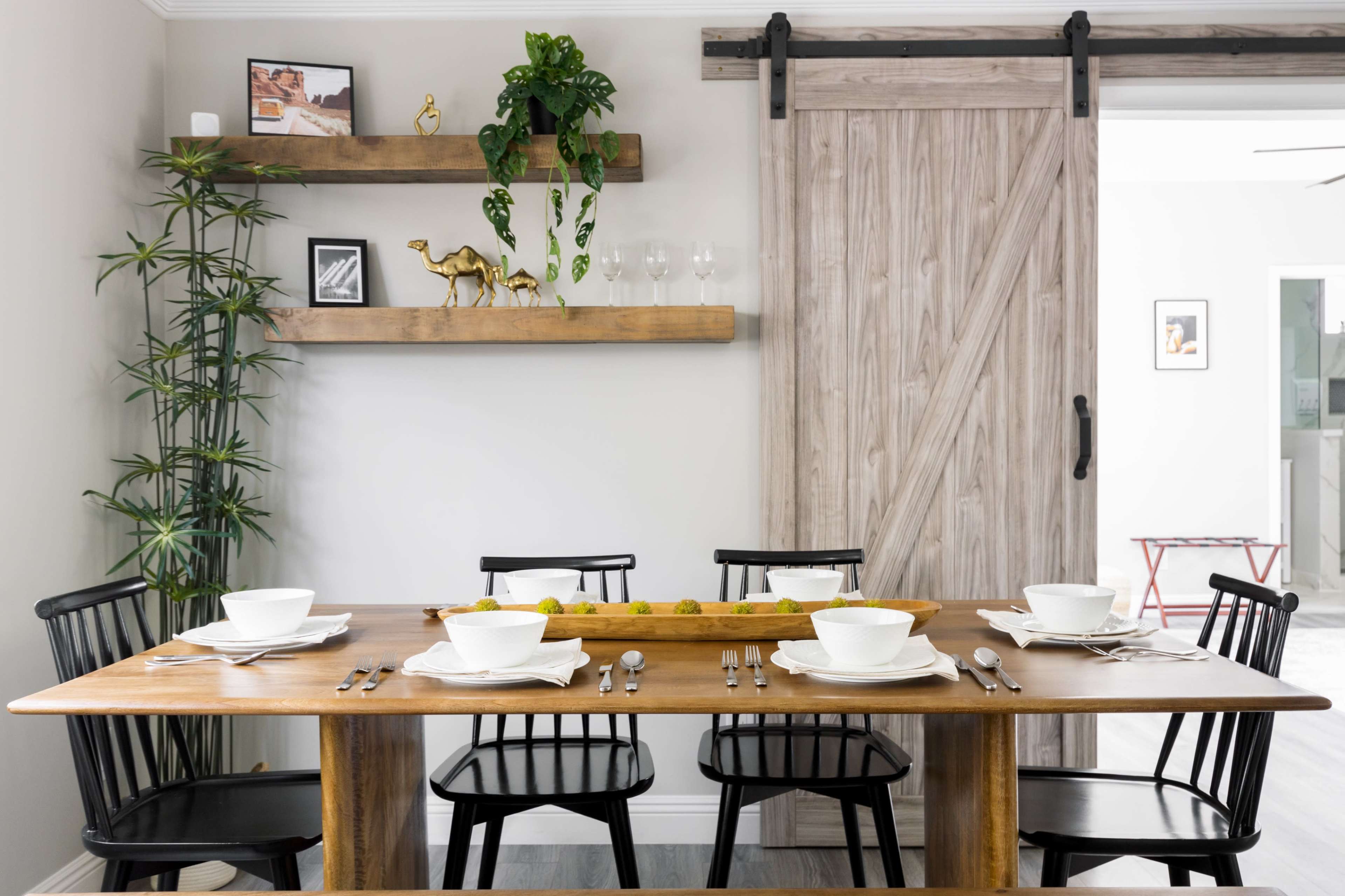 A wooden dining table is set with white bowls and utensils, surrounded by black chairs, alongside a wall featuring shelves with decorative items and a sliding door.