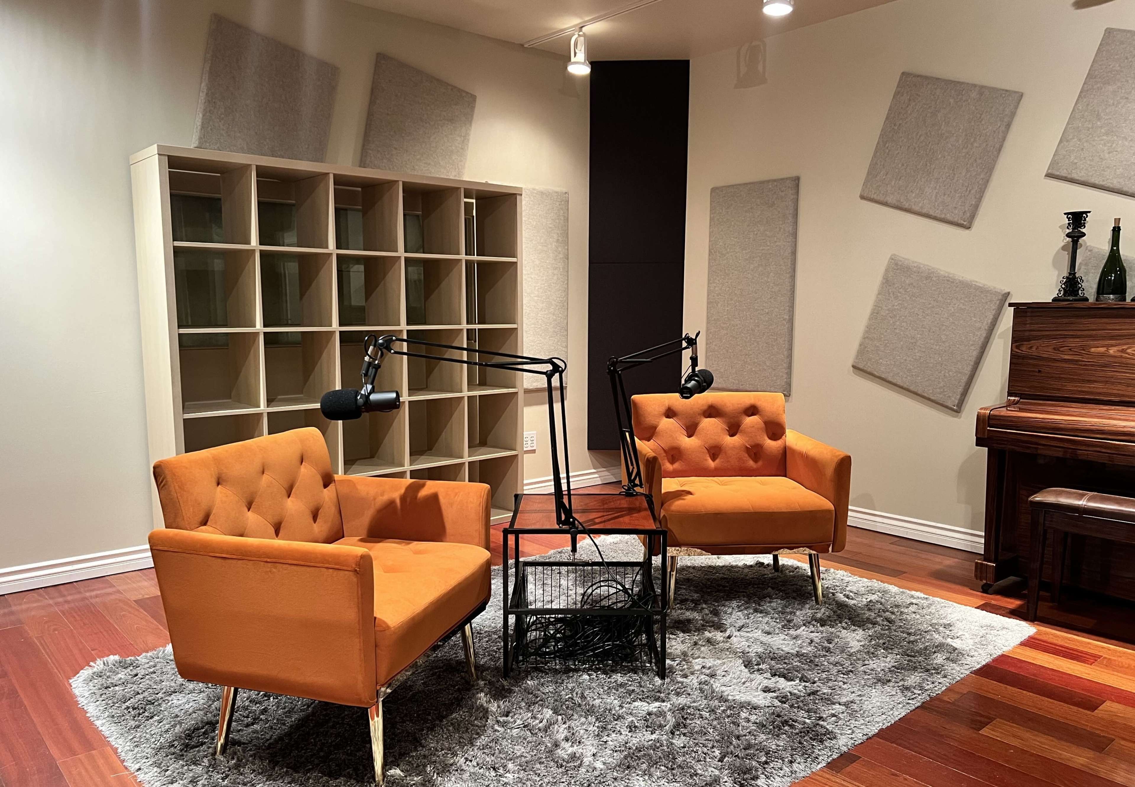 The image shows a podcast recording setup featuring two orange chairs with microphones on stands, situated on a gray rug in a room with acoustic panels on the walls.
