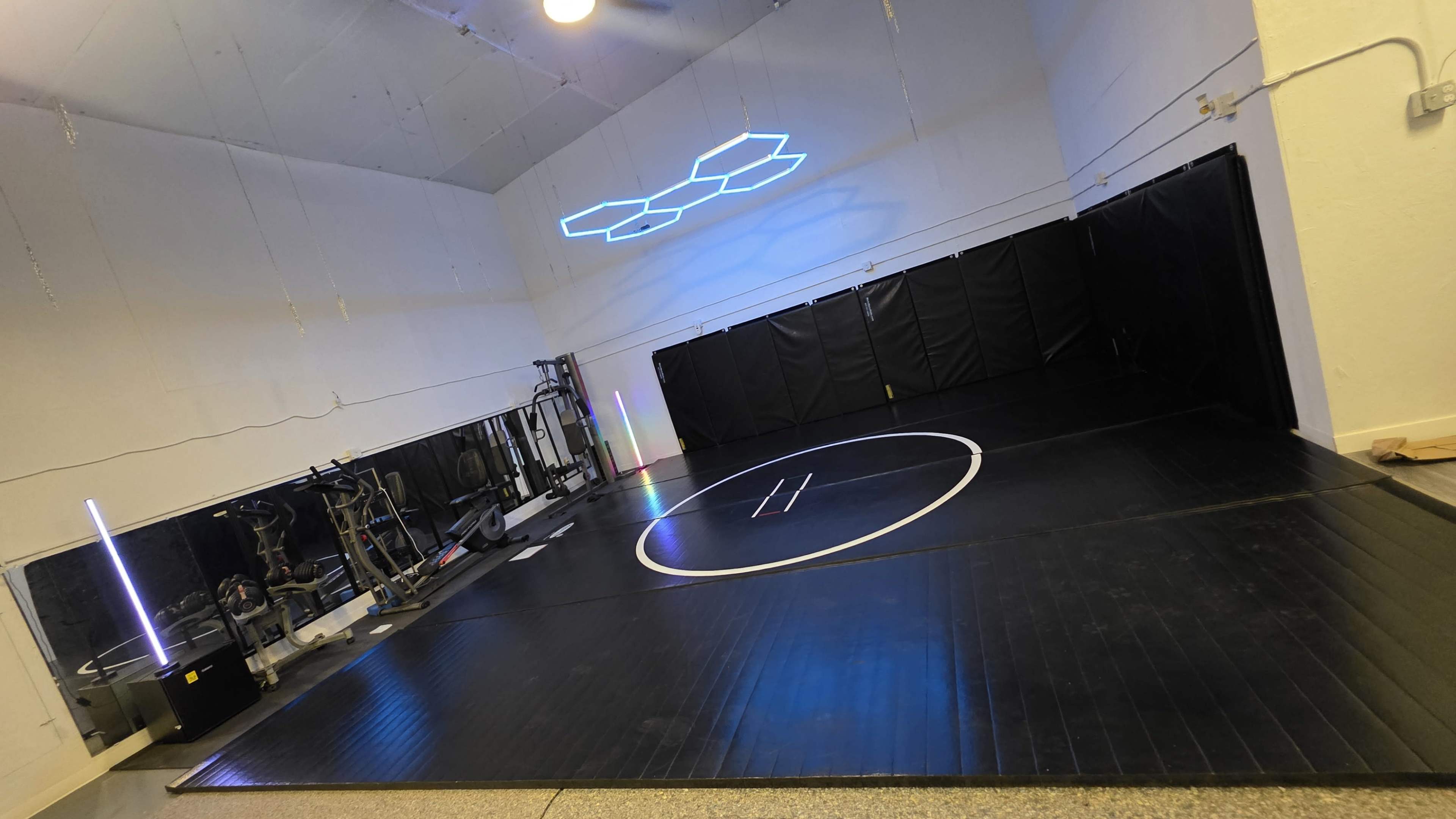 The image shows a spacious training area with a circular wrestling mat, black padded walls, and exercise equipment along one side.