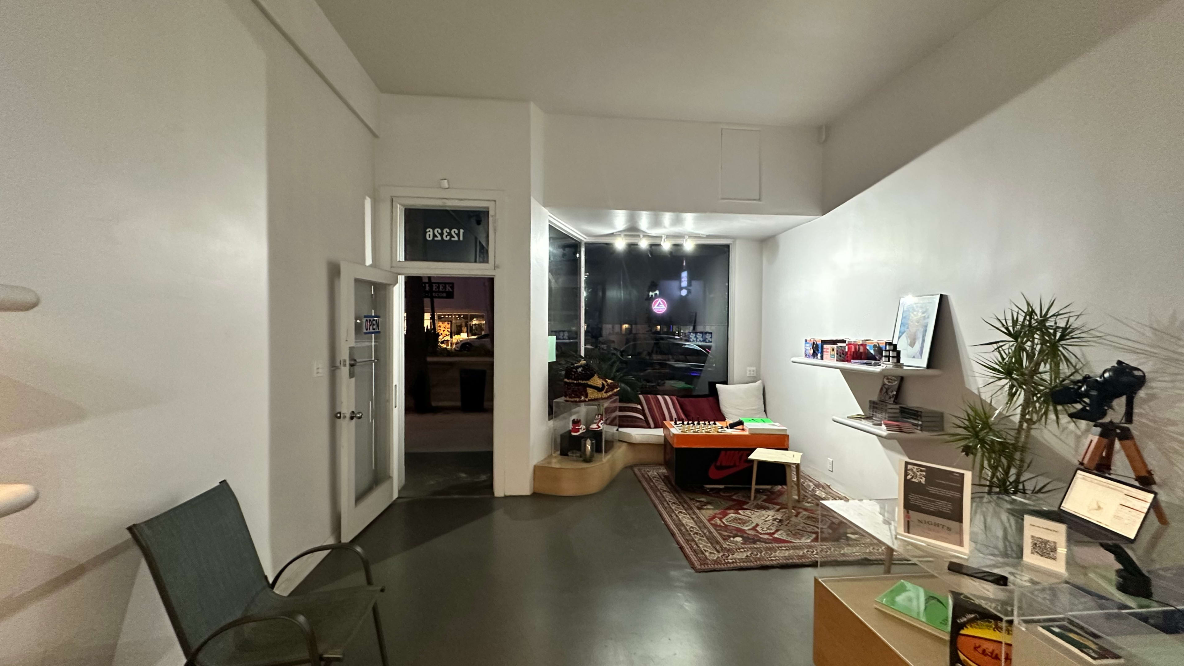 The image shows a modern interior with a minimalist design featuring a seating area, a small table, and a glass door opening to the street outside.