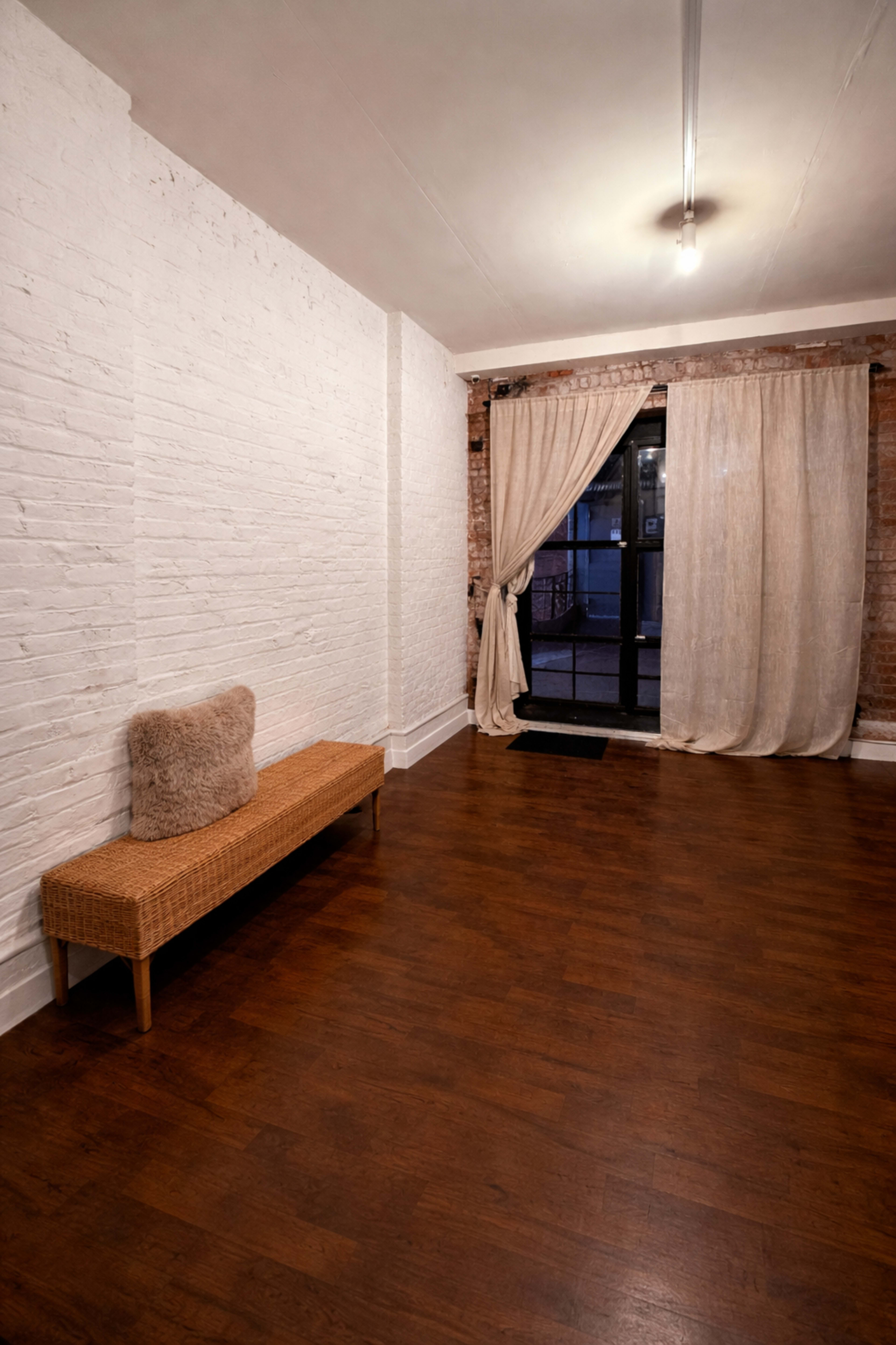 The image shows a sparsely furnished room with a wooden floor, a wicker bench against a white brick wall, and sheer curtains partially covering a large window.