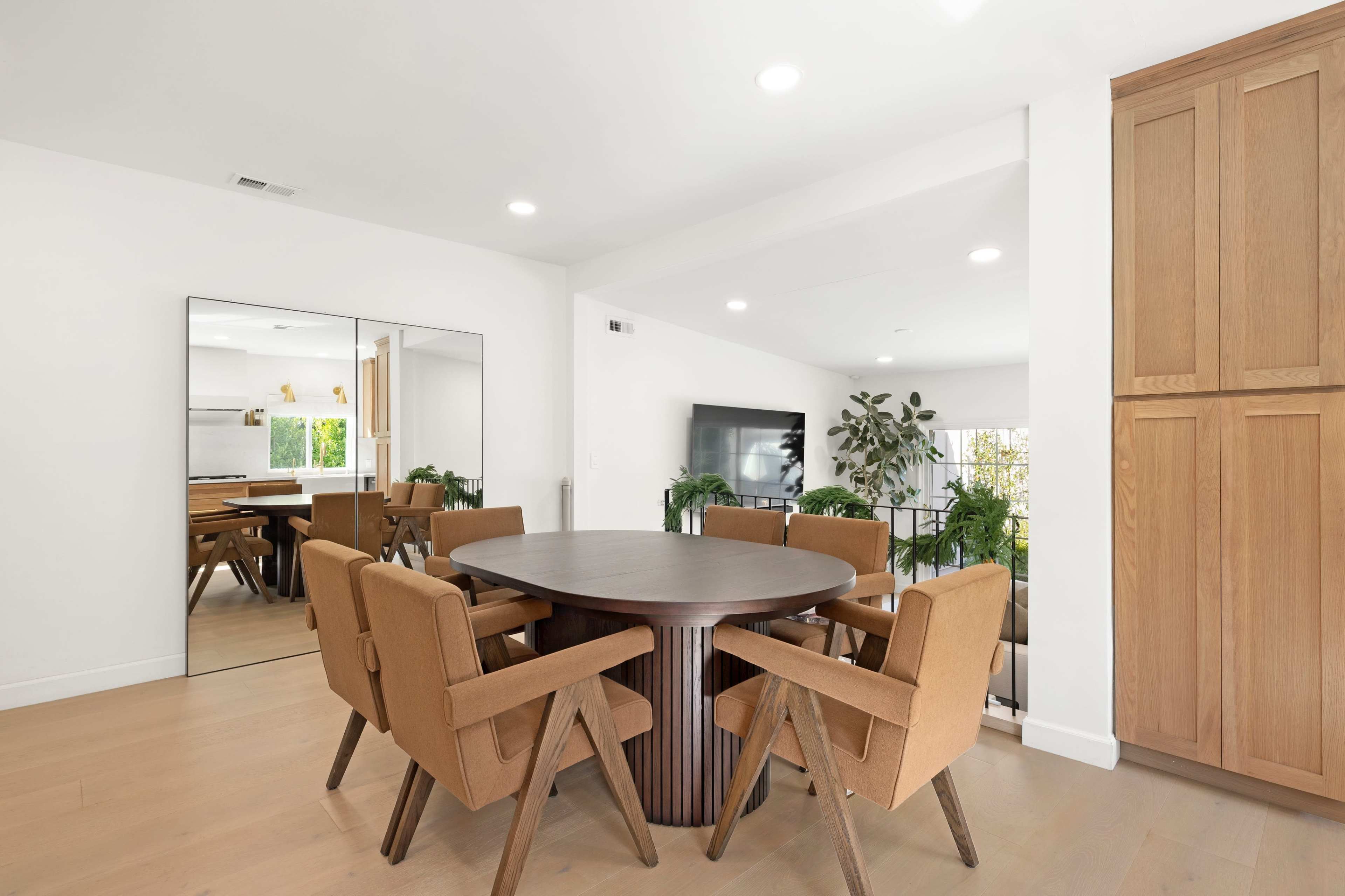 A modern dining area features a circular wooden table surrounded by six upholstered chairs, with a large mirror and plants accentuating the space.