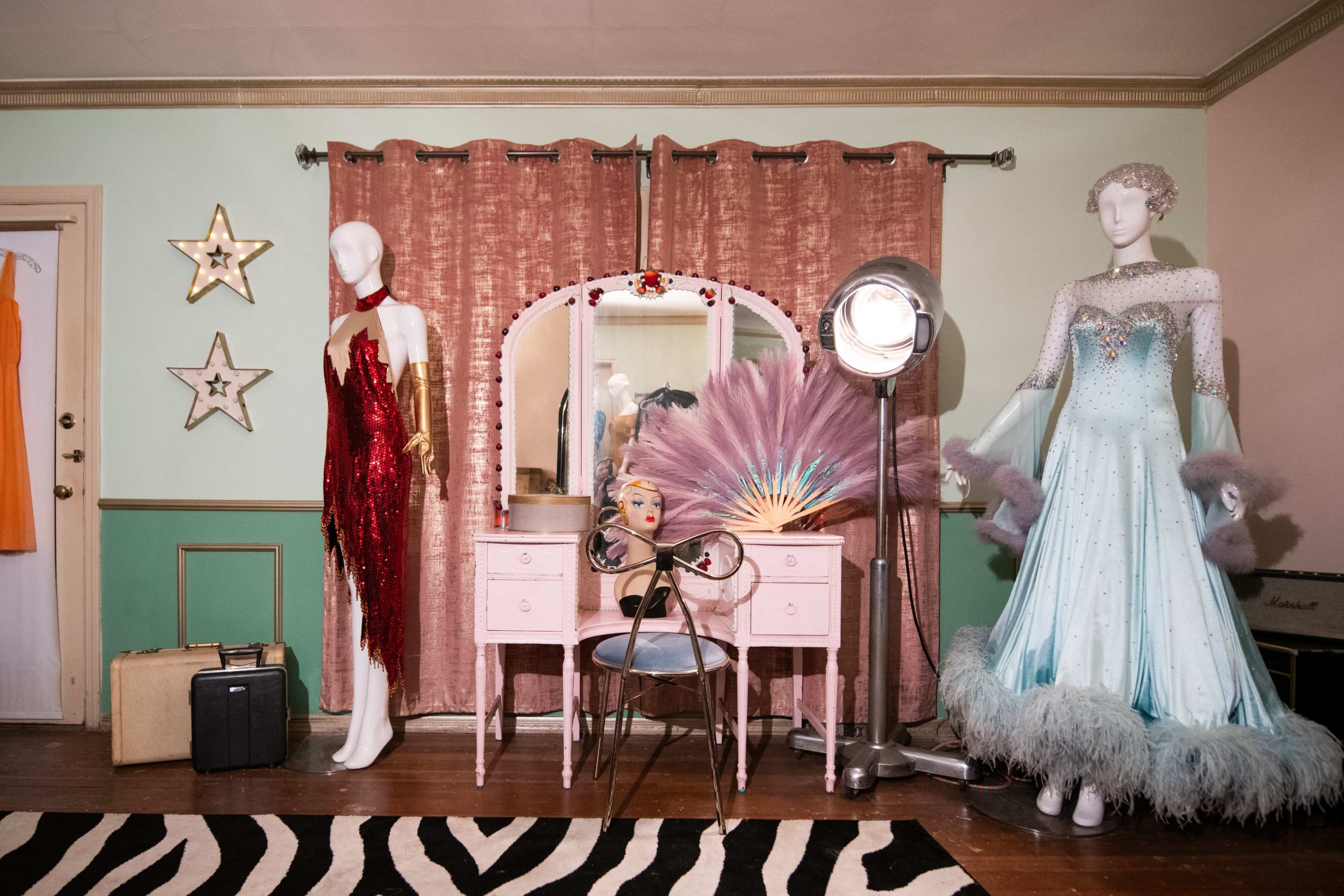 The scene features a dressing room with two mannequins displaying costumes, a vintage vanity with a mirror, and a spotlight, all set against pastel-colored walls and a zebra-striped rug.