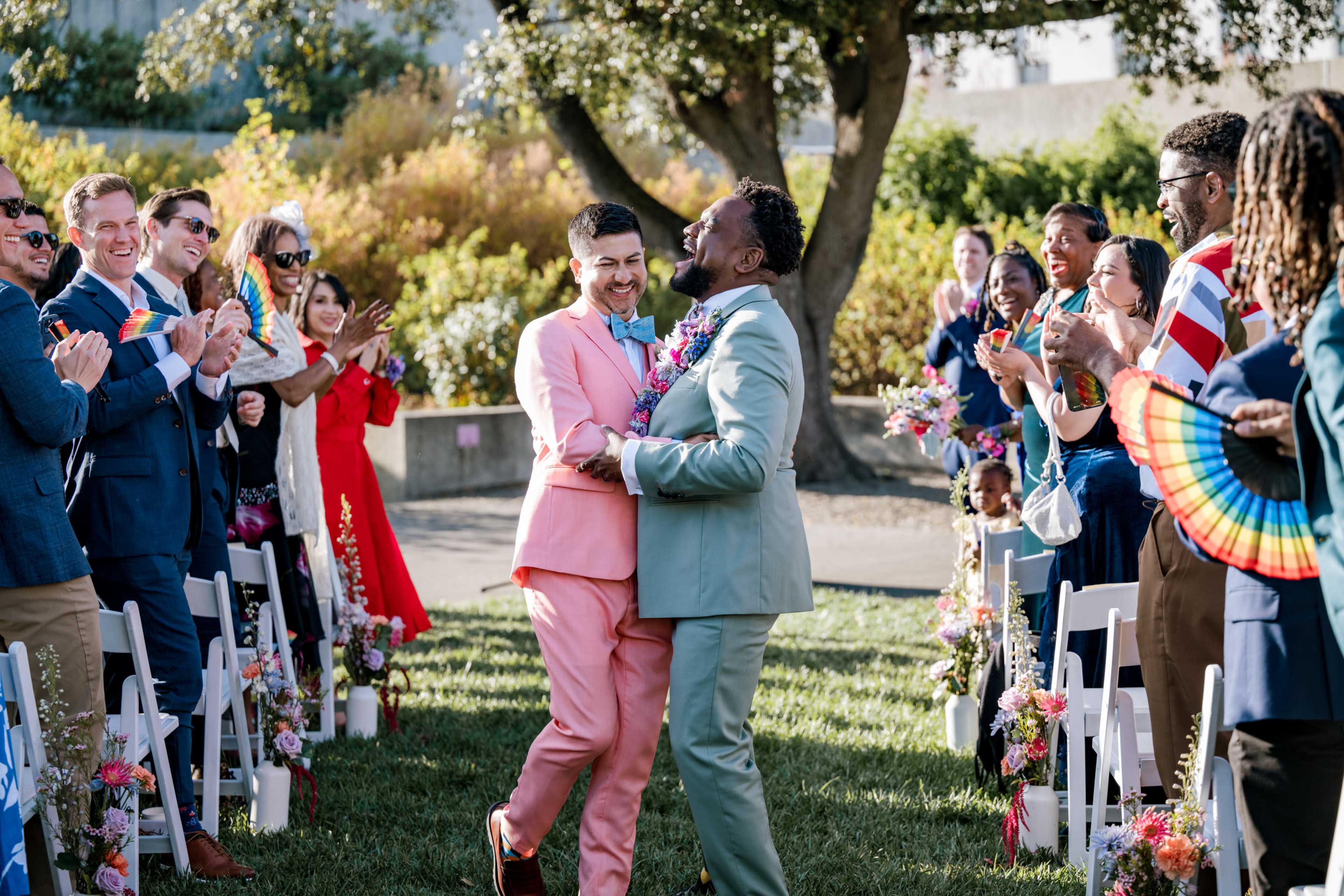 Two men in pastel suits embrace joyfully in an outdoor wedding setting, surrounded by cheerful guests clapping and celebrating.