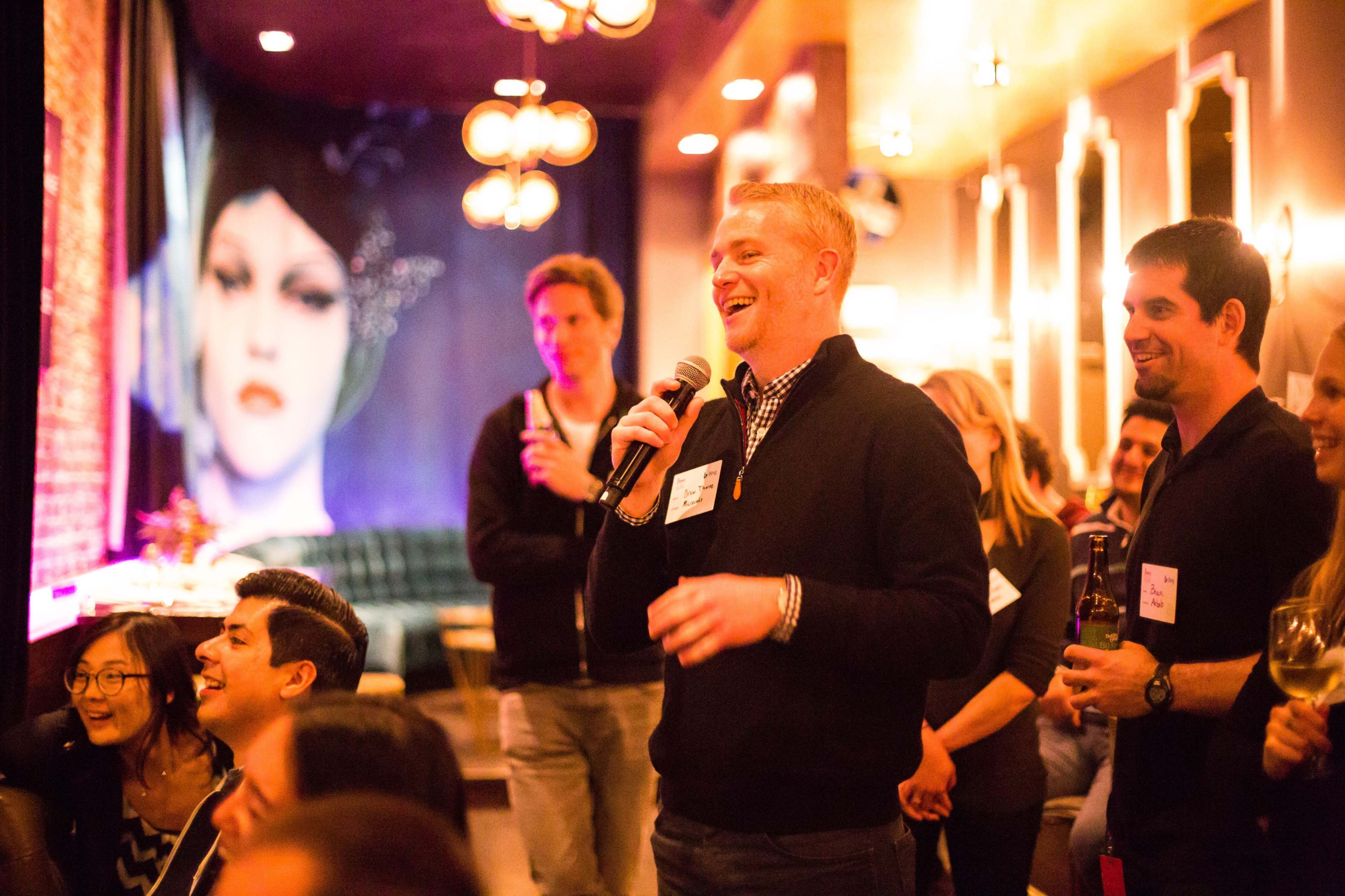 A group of people gathers in a stylish venue, with a man speaking into a microphone while others listen attentively.