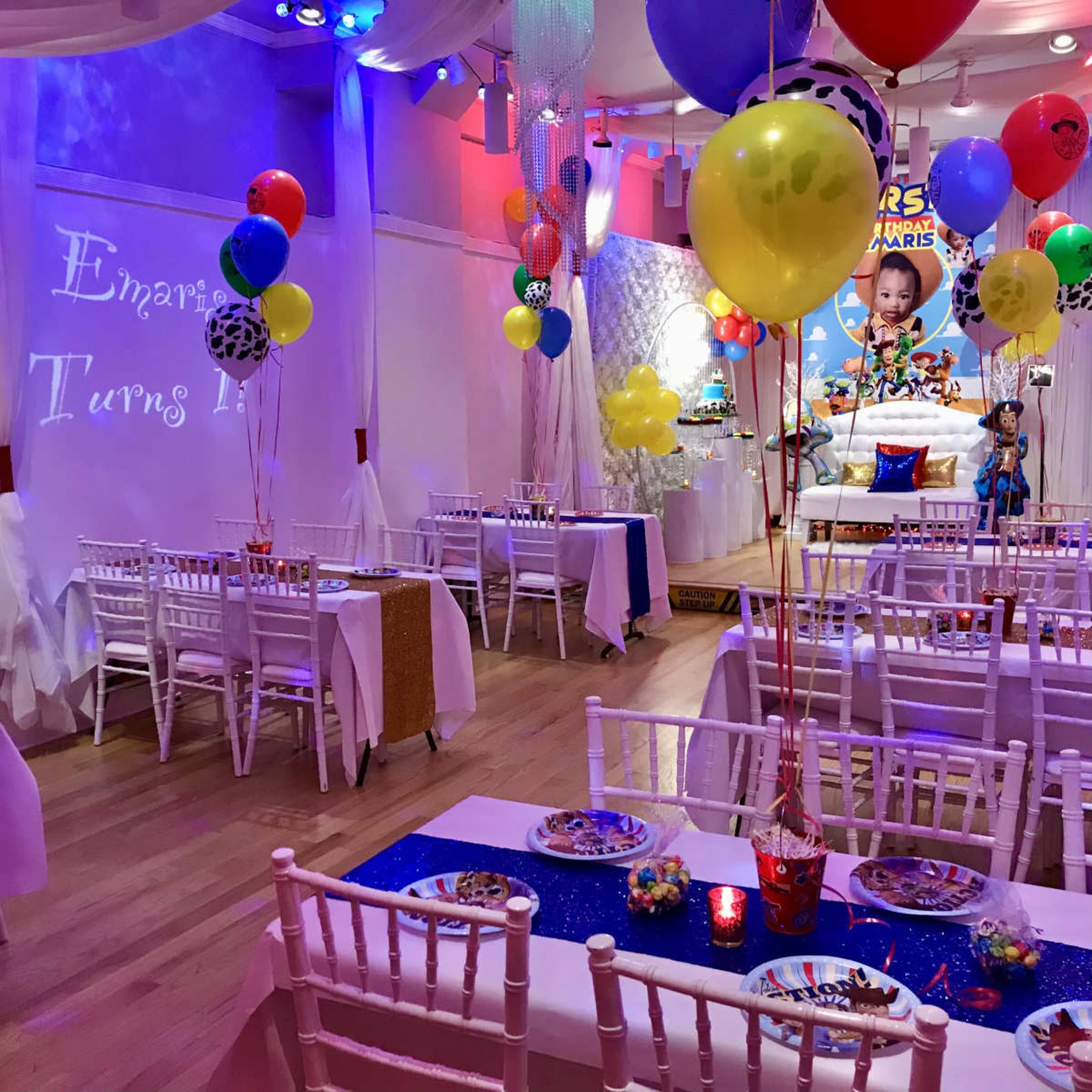 The image shows a decorated party venue with tables set up, colorful balloons, and a birthday theme for a child's celebration.