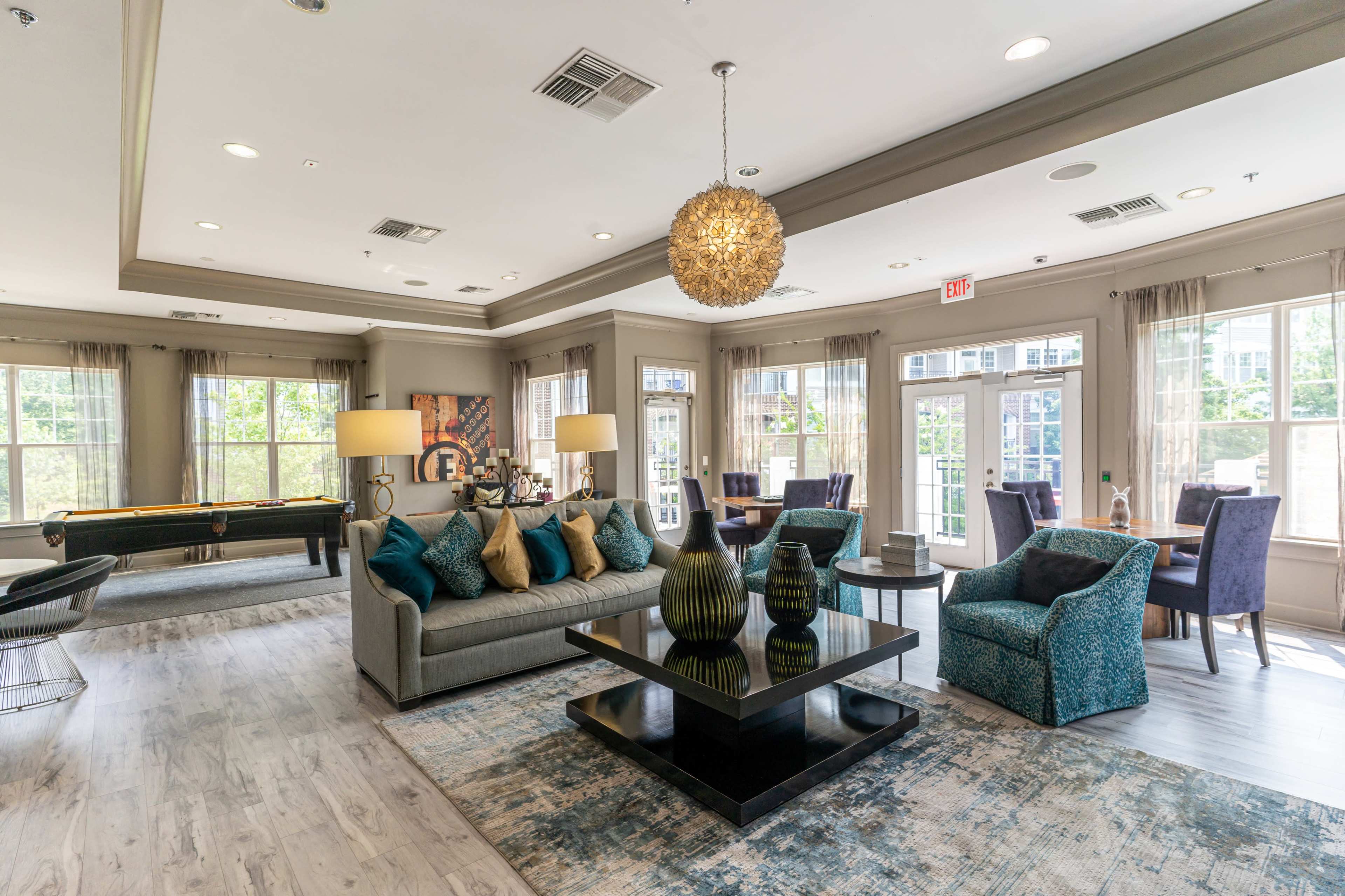 The image shows a stylish lounge area featuring a gray sofa, a black coffee table, decorative vases, and various seating arrangements near large windows.