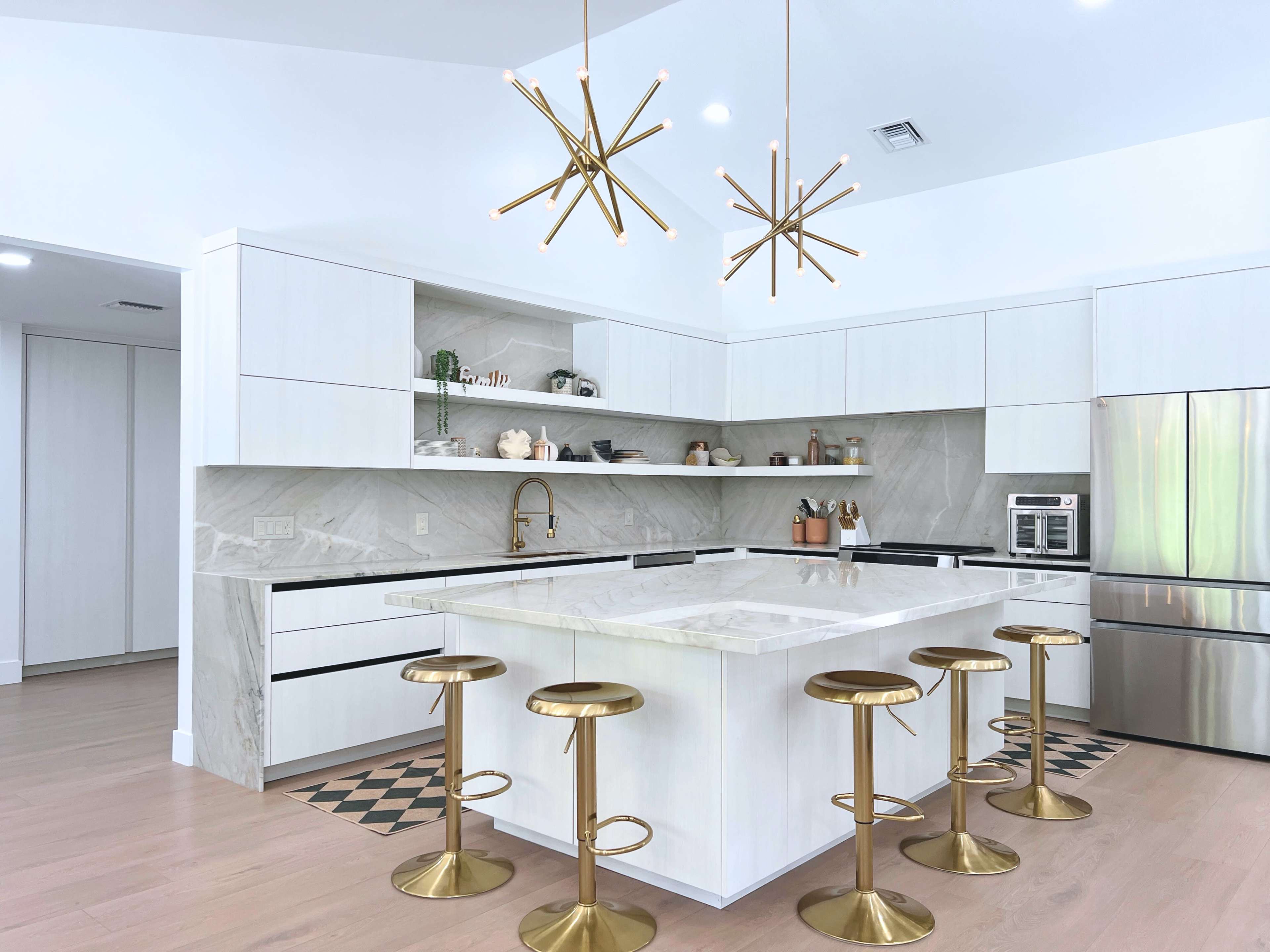 A modern kitchen features a central island with a marble countertop, surrounded by six gold bar stools, and two starburst pendant lights hang above.