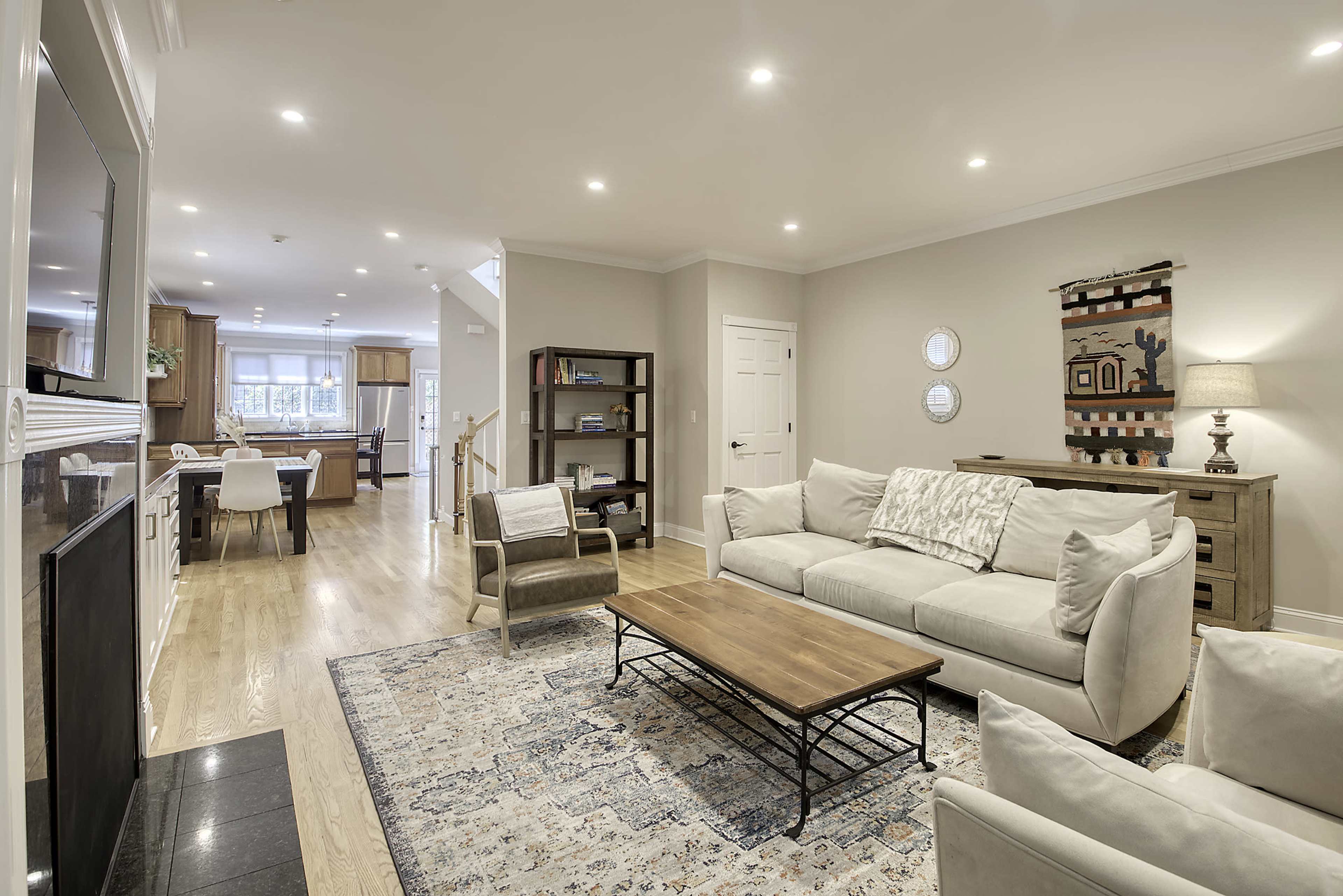 The image shows a bright, modern living room with a light-colored sofa, a wooden coffee table, and a view of an adjacent dining area.