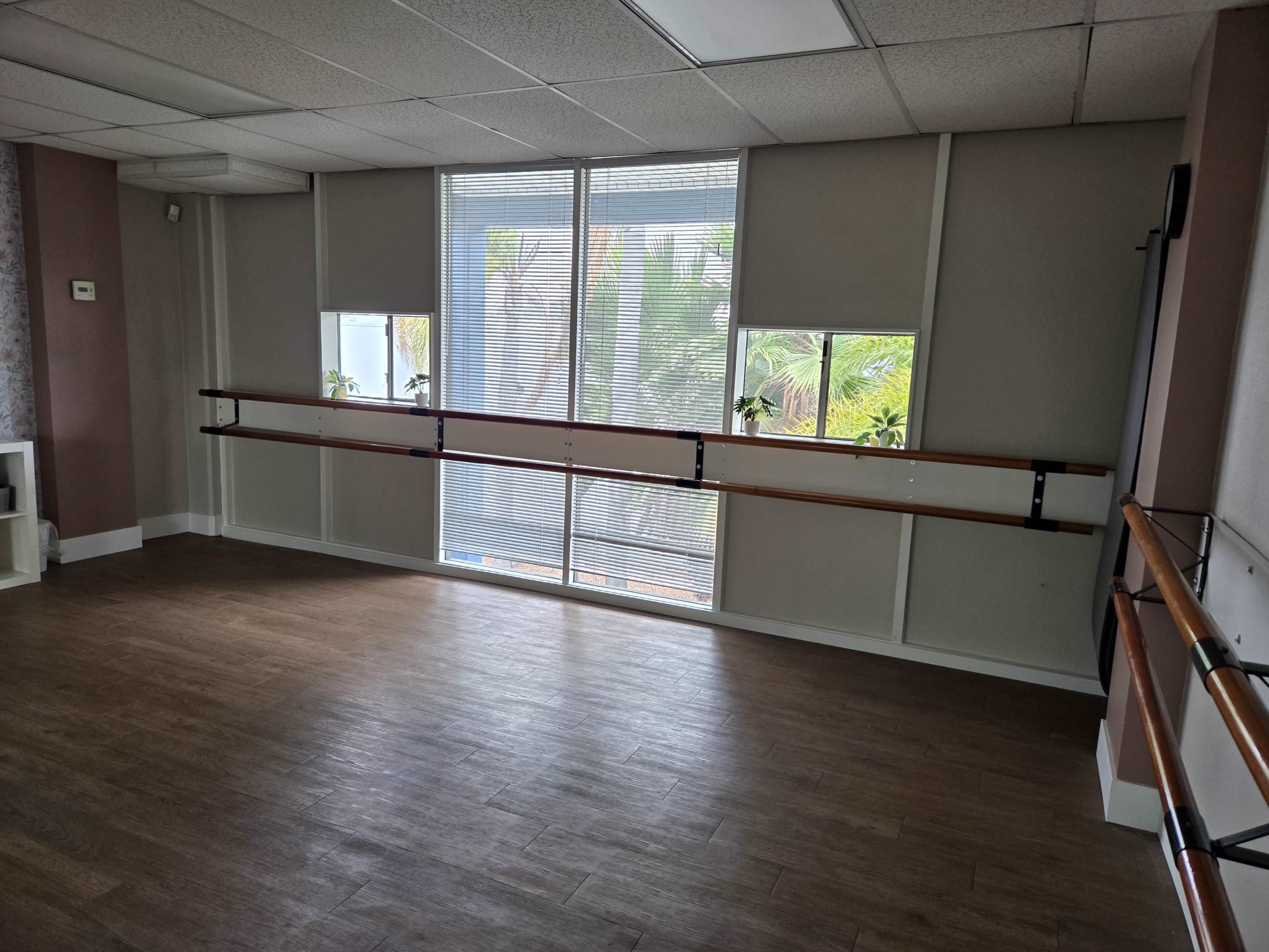 The image shows a dance studio with large windows, hardwood flooring, and a wall-mounted ballet barre along one side.