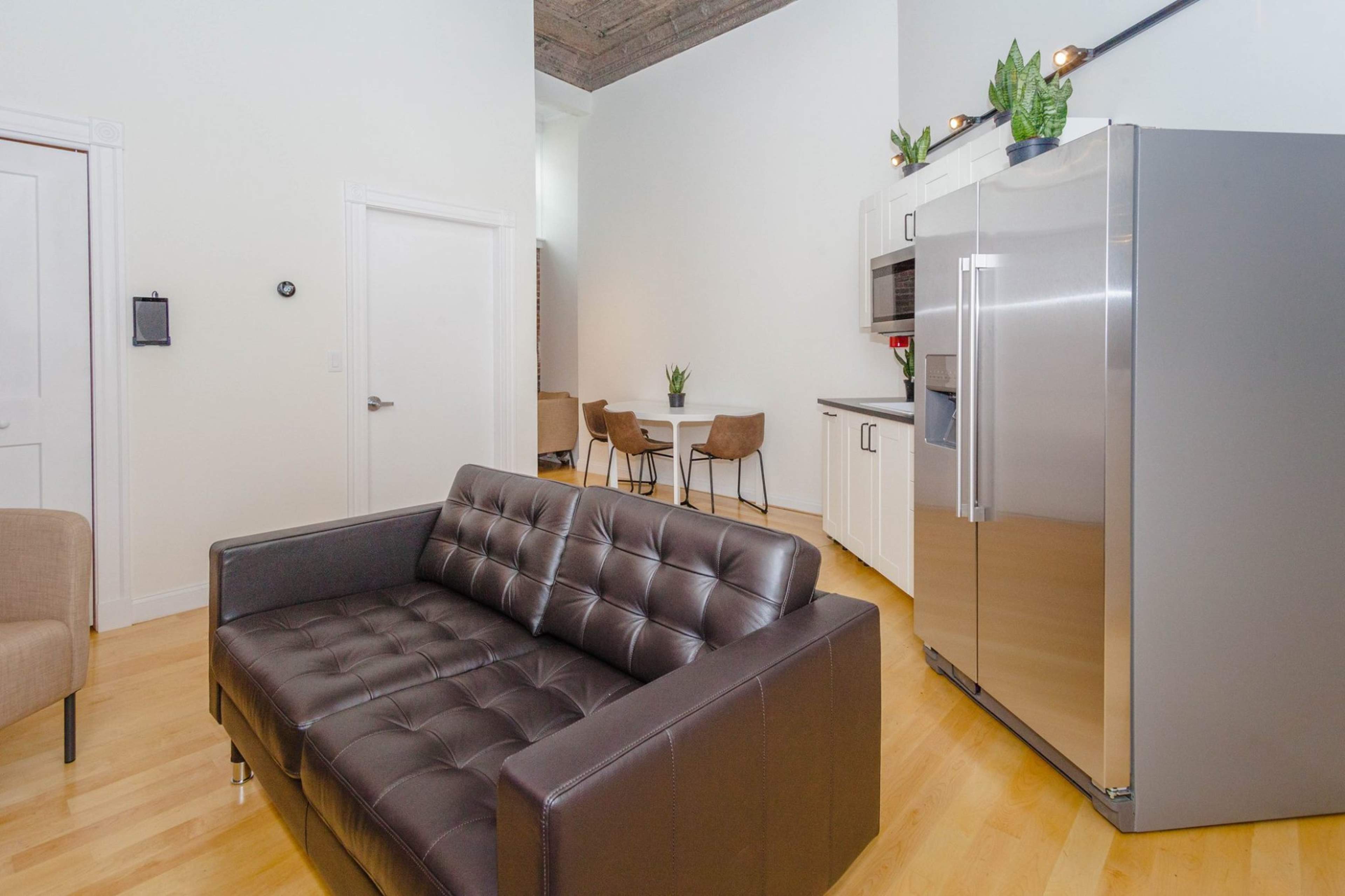 A modern kitchen and seating area feature a black leather couch, stainless steel refrigerator, and a dining set in the background.