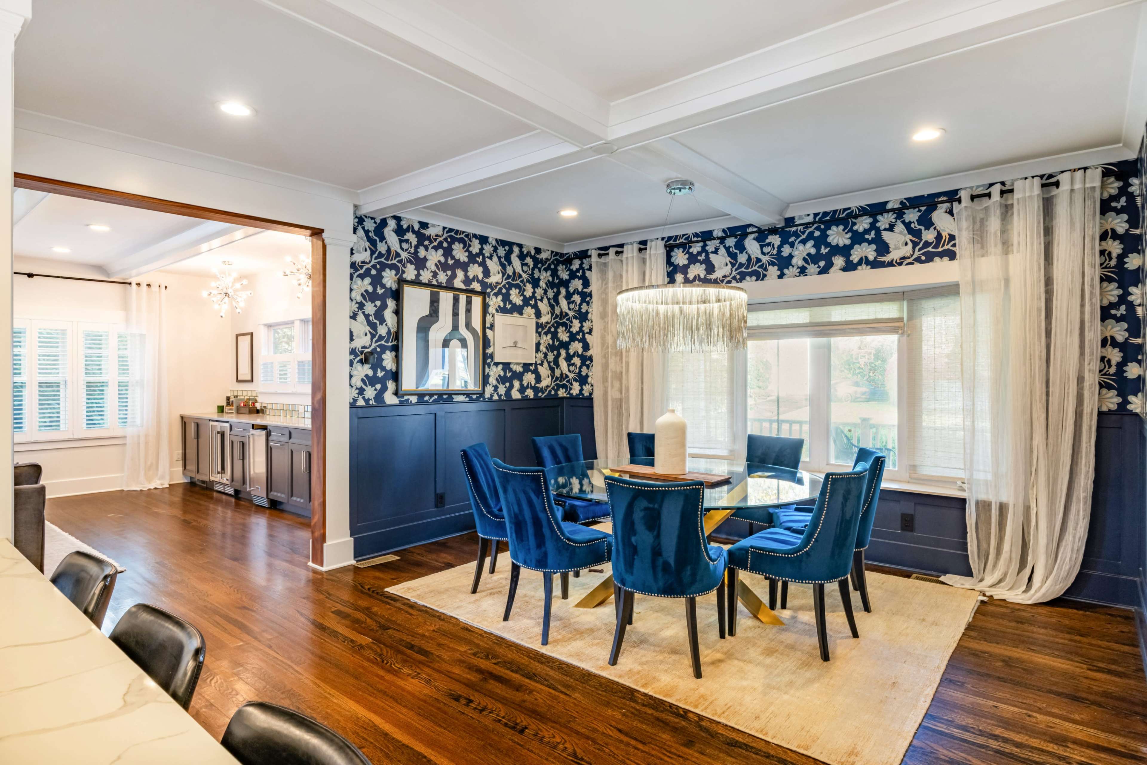 The image shows a dining area featuring a round table surrounded by blue chairs, set against a floral wallpaper and illuminated by a large pendant light.