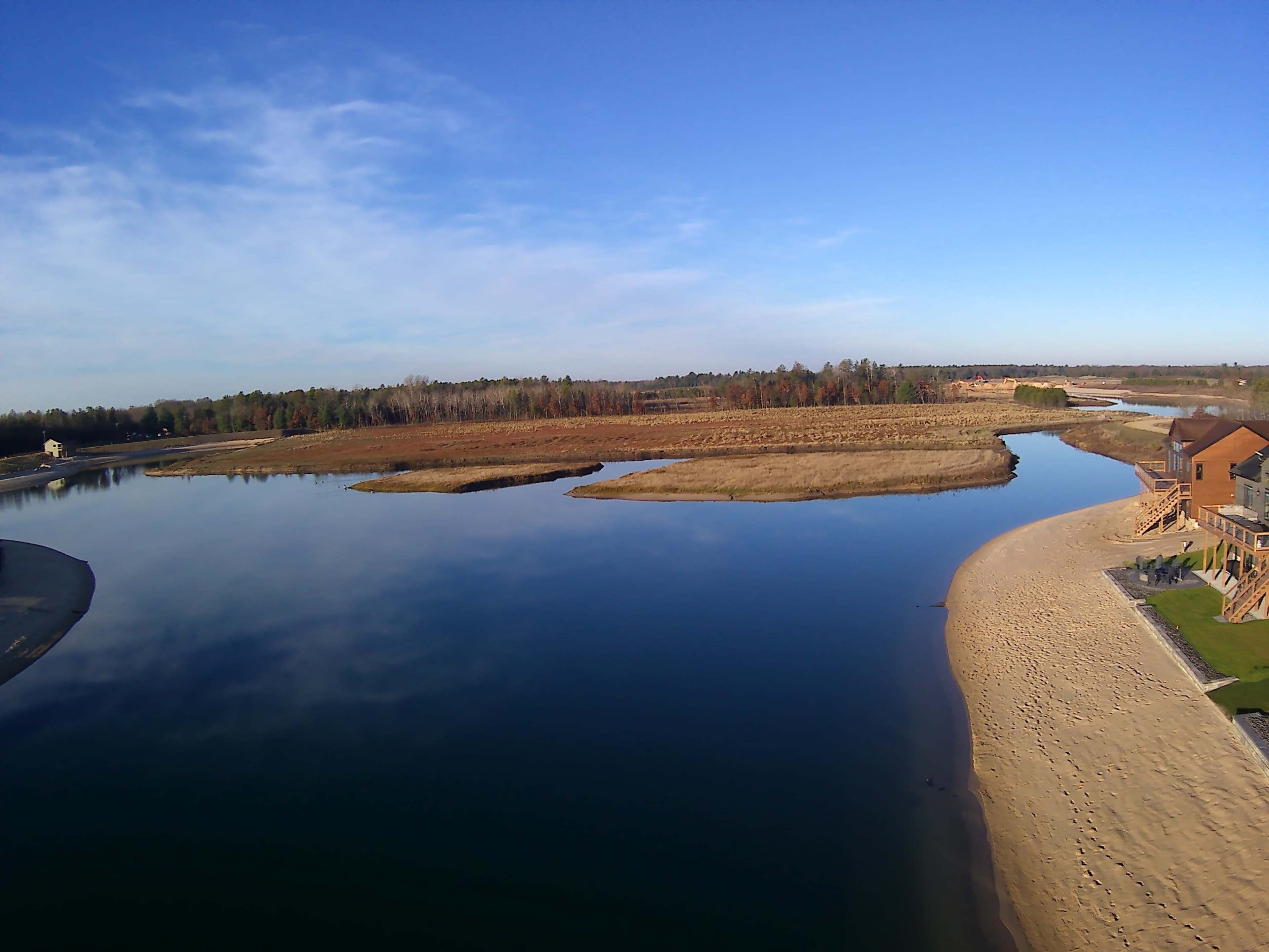 A calm river bends through a landscape with grassy banks and a sandy shore, bordered by trees in the distance under a clear blue sky.