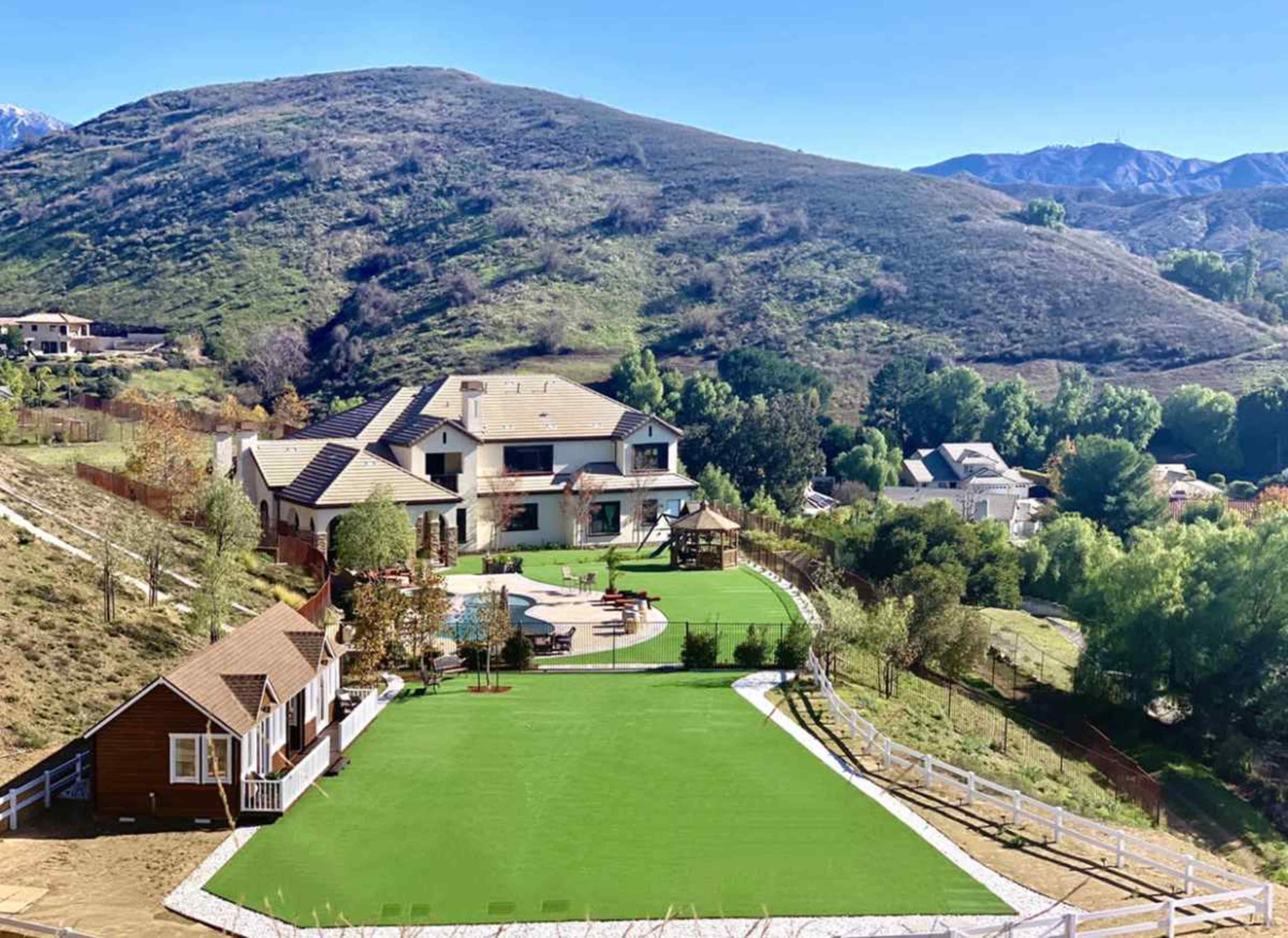 The image shows a large residential property with a green lawn, a swimming pool, and a playground, surrounded by hills and trees.