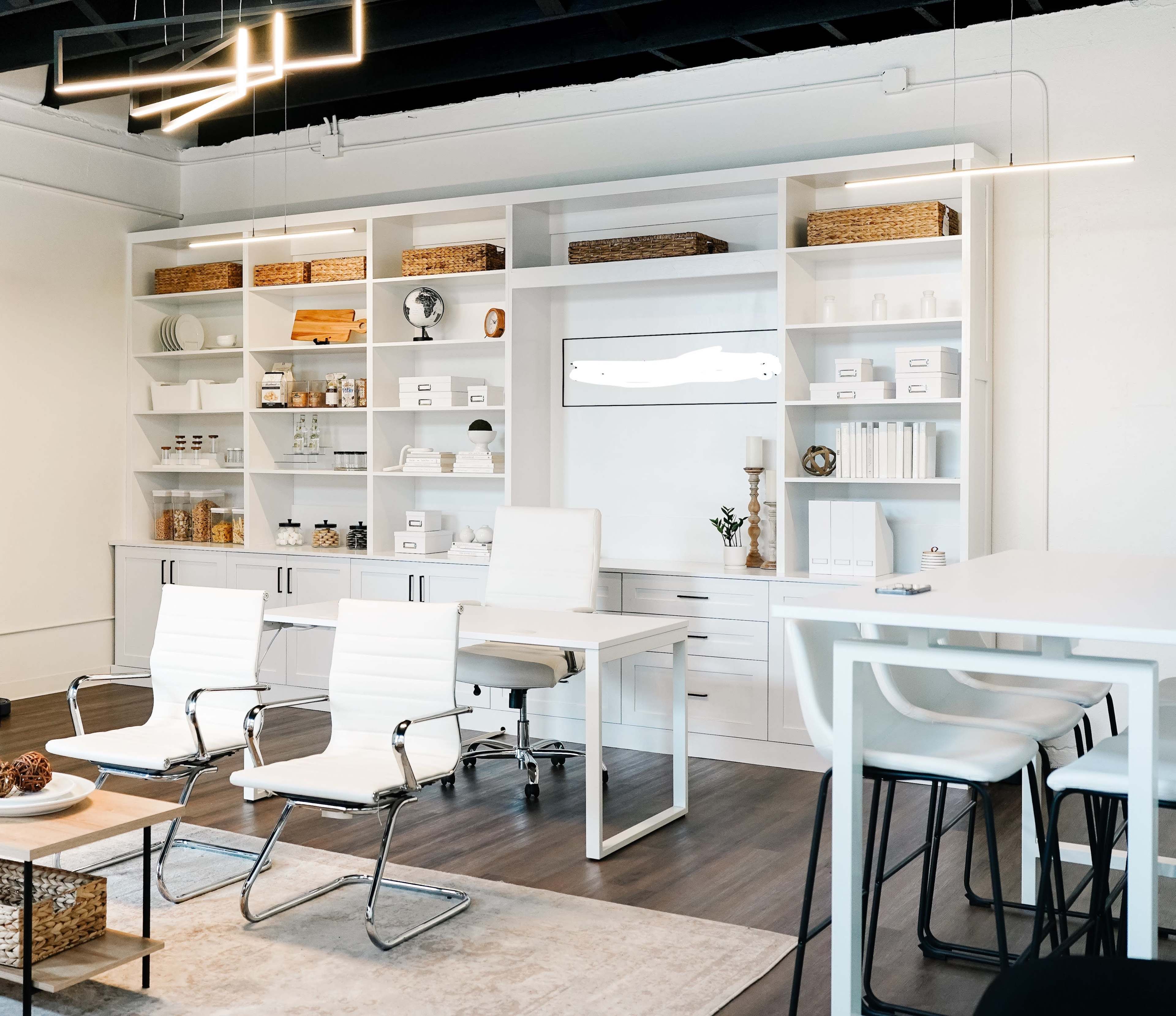 The image depicts a modern office interior featuring a sleek desk, two white chairs, and shelving organized with various decorative and storage items.