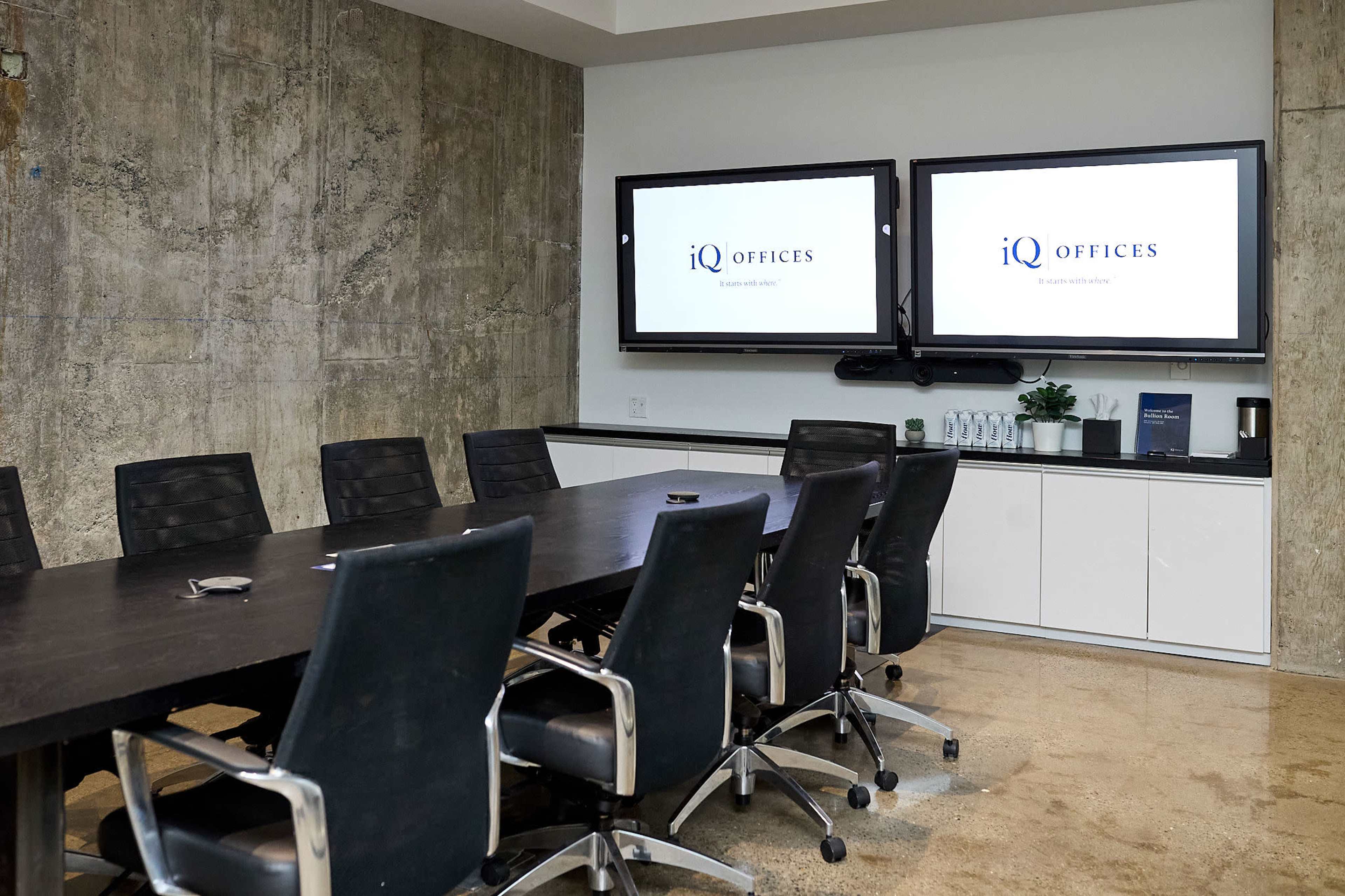 A modern conference room features a long dark table surrounded by black ergonomic chairs, with two screens mounted on a concrete wall displaying the "iQ Offices" logo.